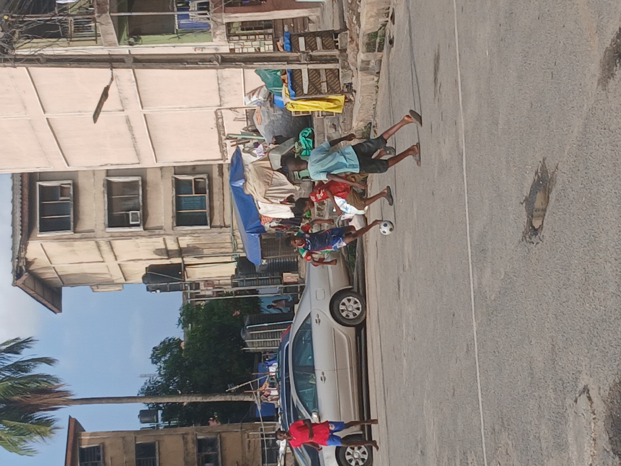 Children are playing soccer in the street in Lagos, Nigeria. Four boys are mid-play, interacting with the soccer ball. A silver sedan car is parked on the side of the road. Buildings are visible in the background, some with balconies and clotheslines. A makeshift awning or tent-like structure is visible near one of the buildings. A palm tree is to the left of the image. The street has visible wear and tear. The sky is partly cloudy.