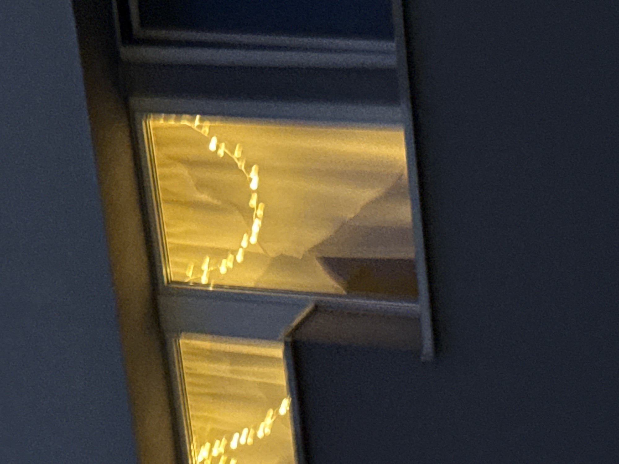 An outdoor, close-up, eye-level shot captures a dark blue building facade with two windows at dusk. The left window is framed by a light-colored curtain, and both windows are adorned with string lights that emit a warm yellow glow. The top window frame is illuminated by the warm light of the string lights. The overall lighting suggests the scene is captured at twilight or night.