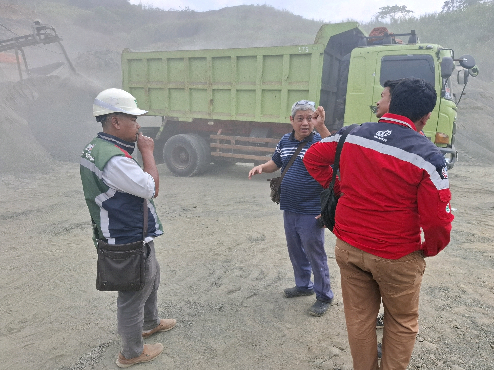 The image depicts an outdoor scene in Cijangkar, Indonesia, likely a construction or industrial site given the presence of a large green dump truck. In the foreground, three men are interacting. On the left, a man wearing a white hard hat and a green and white vest over a white shirt is facing the right, appearing to speak or gesture with his right hand near his mouth. He has a dark shoulder bag. In the center, a man wearing a blue and white striped shirt and glasses on his head is facing the left, gesturing with his right hand raised as if explaining something. He is also wearing a dark shoulder bag. On the right, a man in a red and dark blue jacket and brown pants is facing away from the camera, towards the dump truck. The dump truck is light green and appears to be partially loaded with gravel or sand, with a conveyor belt system visible in the background on the left, also dispensing material. The ground is a mix of dirt and gravel, and a light dust or mist is present in the air, obscuring some of the background details. The sky is overcast.