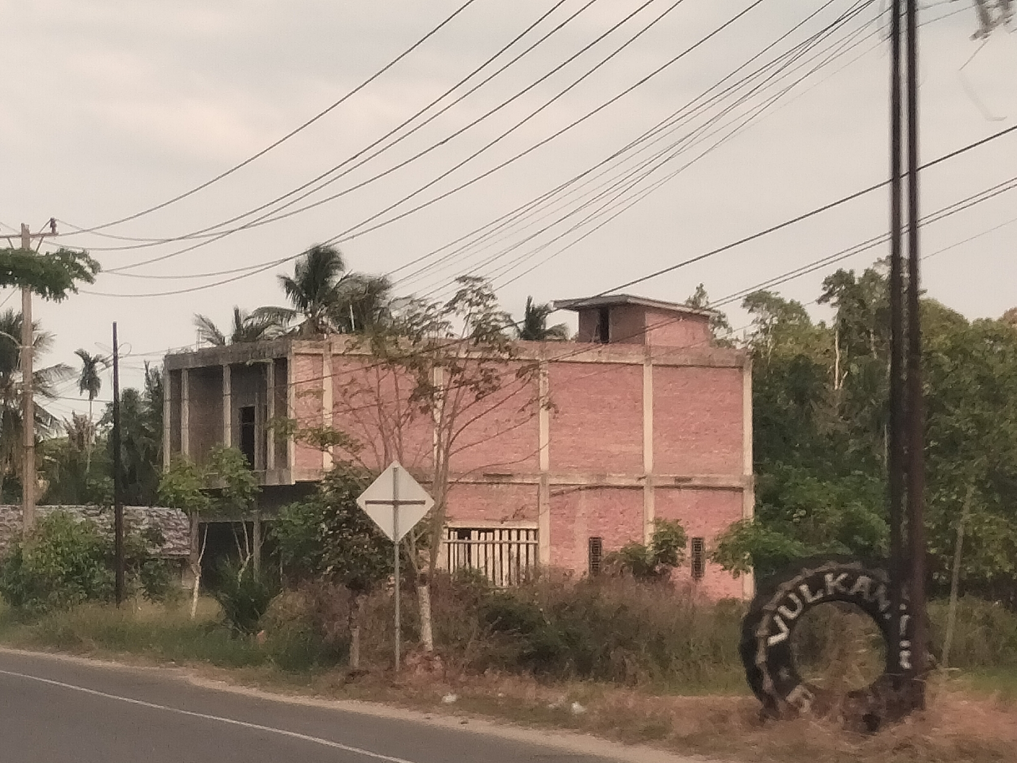 The image shows an unfinished, two-story brick building in Teupinbatee, Indonesia. The building has open column supports, suggesting an incomplete construction phase. A road sign and a tire with the word "VULKANISIR" painted on it are situated along the roadside. Overgrown vegetation surrounds the building, indicating a period of neglect. Power lines run overhead. The sky is overcast.