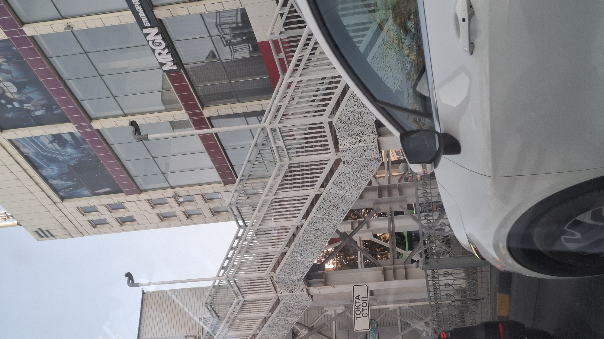 A street-level, eye-level, medium shot captures a white car in the foreground, facing right, with its rear portion partially visible on the left. The car's right side, including the door handle and side mirror, is clearly in view. Behind the car, a metal fence runs horizontally across the frame. To the left of the fence, a pedestrian overpass is prominent, with white stairs and railings. A rectangular sign with Cyrillic text, reading "ТОКТА СТОП" (meaning "Stop"), is attached to the overpass. In the background, a modern building with large glass windows and a facade adorned with posters is visible. The building has a sign that reads "MRGN БИЛЬЯРД" (meaning "MRGN Billiards"). The sky above is clear and bright. The overall scene depicts an urban environment, likely in the city of Shymkent, Kazakhstan.