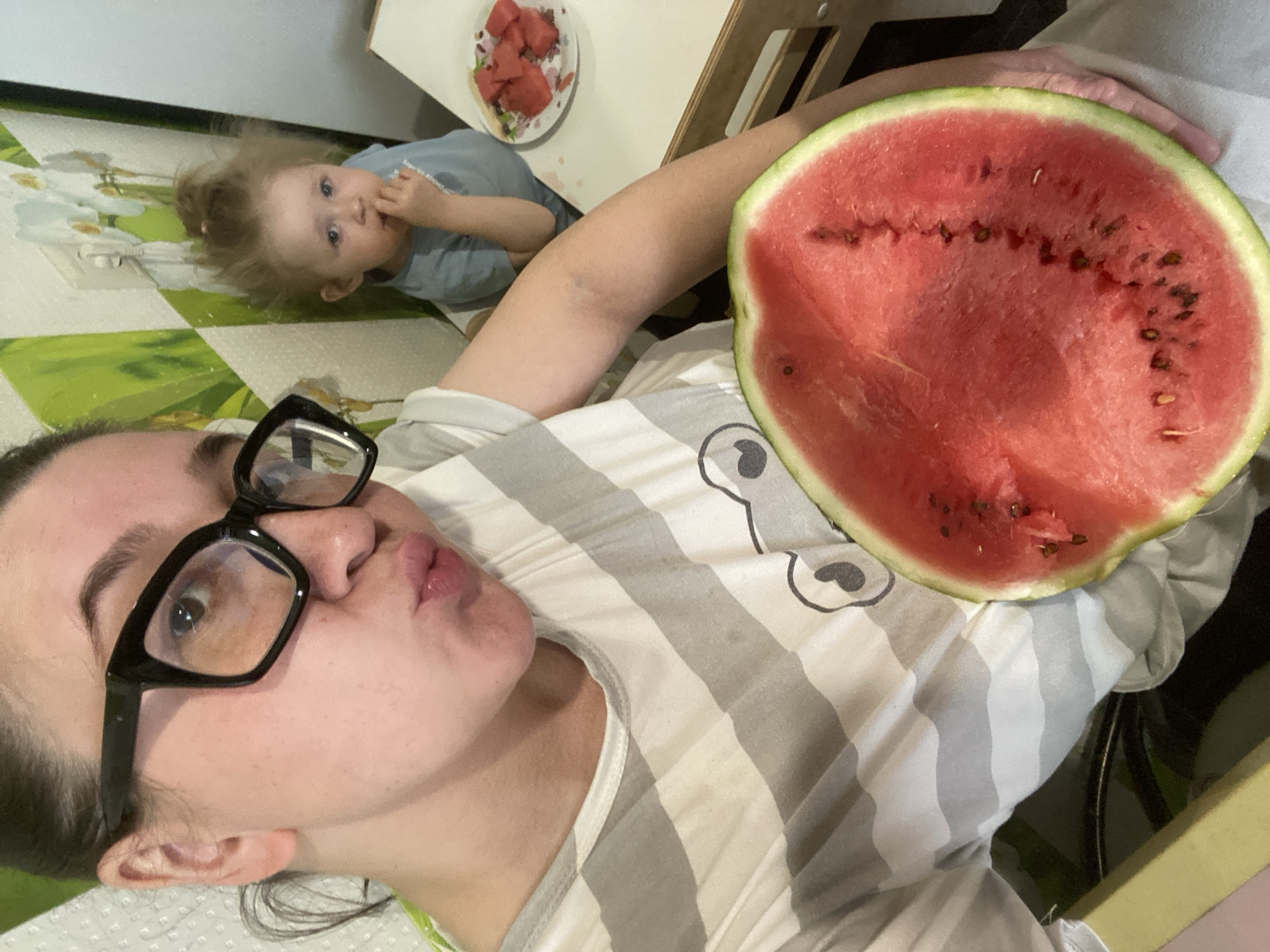 In an indoor setting, potentially a kitchen or dining area, a woman with dark hair pulled back and wearing large black-rimmed glasses takes a selfie. She is puckering her lips and holding a large slice of watermelon. She is wearing a grey and white striped t-shirt with a graphic on it. To her right, a young child with blond hair in a messy bun is seated at a table, eating watermelon. The child is wearing a light blue shirt. A plate of chopped watermelon is visible on the table in front of the child. The background features a wall with a green and white floral pattern. Based on the provided information, the location is the city of Chaikovsky, Russia.