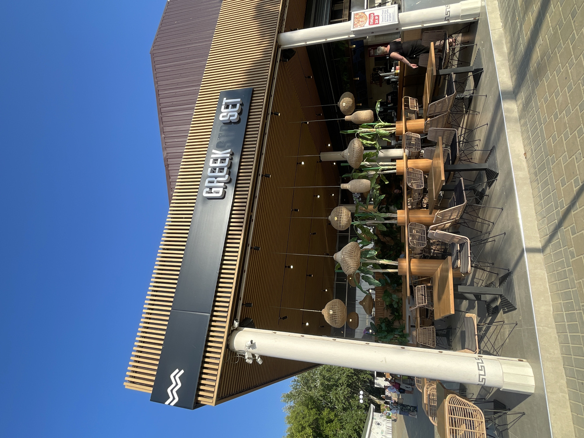 The media file shows an outdoor seating area of a restaurant with a prominent sign that reads "GREEK SET." The restaurant has a modern facade with wooden slatted accents and a black sign with white lettering. Woven pendant lights hang over several tables and chairs, suggesting an evening setting or a desire for ambient lighting. Lush green plants are interspersed throughout the seating area, adding to the ambiance.
In the background, a person appears to be working behind a counter, possibly serving customers. Other tables are occupied by patrons, indicating a lively atmosphere. The ground is paved with light-colored bricks, and a sidewalk is visible. A clear blue sky suggests good weather and likely daytime. The overall impression is a relaxed and inviting dining environment.
A sign on the wall of the building displays text in Cyrillic, which translates to "pizza for you" and further details about the pizza. The setting appears to be a street or promenade in Anapa, Russia, a coastal city known for its resorts. The time of day appears to be late afternoon or early evening, given the lighting and the shadows cast. The mood is one of casual enjoyment and hospitality.
