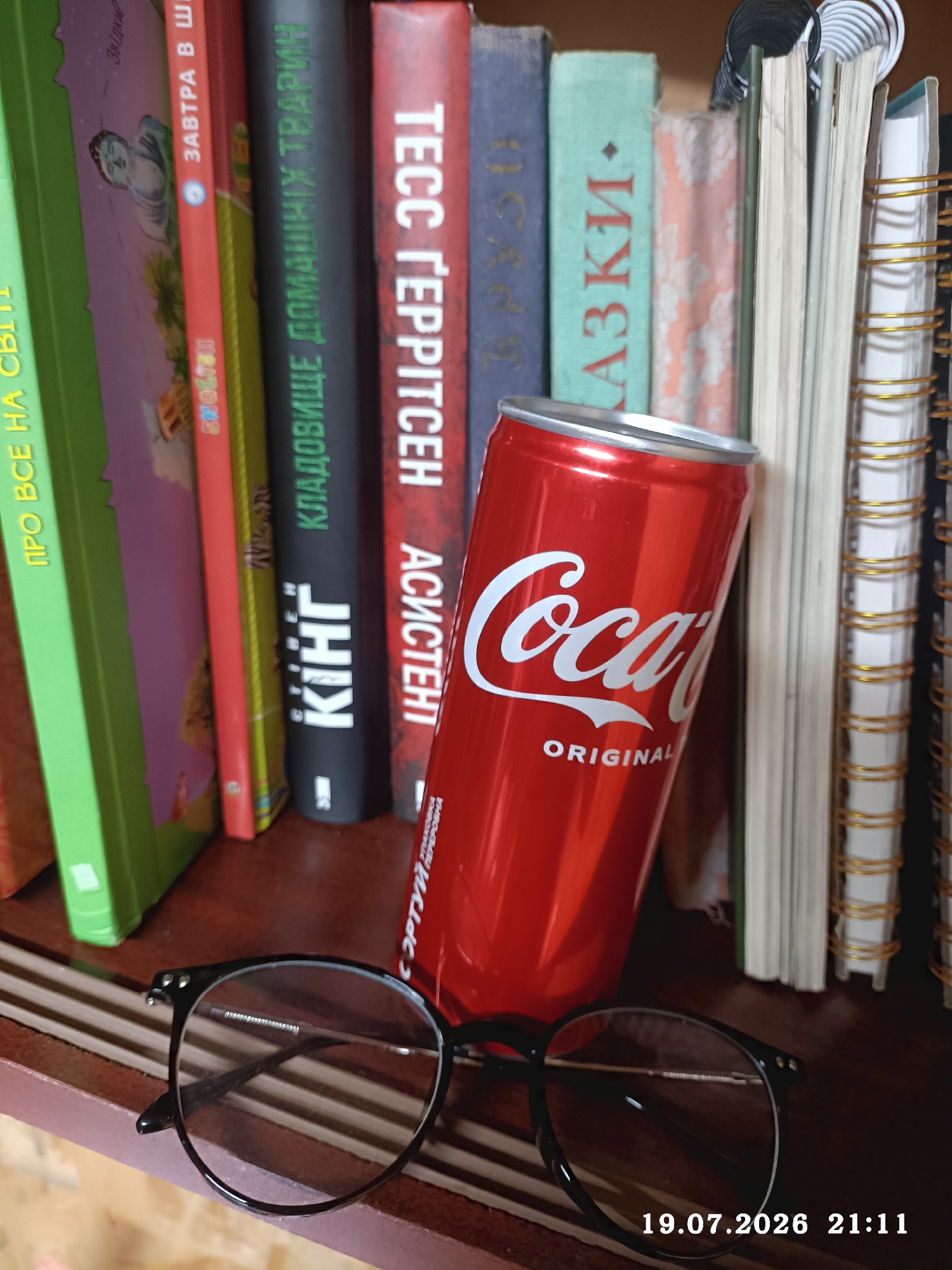 An indoor, eye-level shot captures a shelf with several books and a Coca-Cola can, with a pair of black-rimmed glasses in the foreground. The books are of varying sizes and colors, with visible titles in Cyrillic script. One book has a green cover with yellow text, another is red with white text, and a third is black with white text. A red Coca-Cola can with the iconic white logo and the word "ORIGINAL" printed below it is placed leaning against the books. The glasses in the foreground are simple, black-rimmed spectacles. The background appears to be a wooden shelf. A timestamp in the bottom right corner reads "19.07.2026 21:11". The scene does not provide any specific details to identify its location within the city of Radisne, Ukraine.
