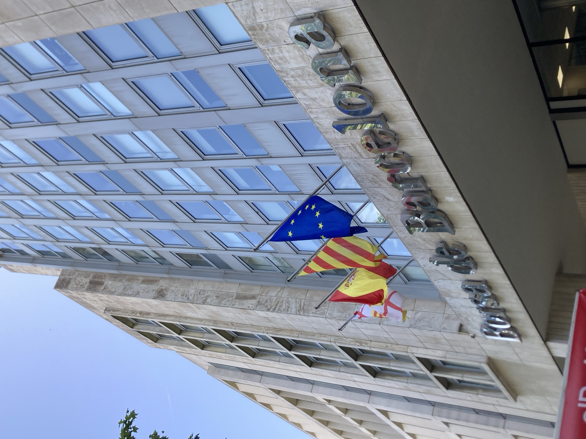 This media file is a low-angle shot of a modern building facade in Barcelona, Spain. The building features a grid of numerous windows reflecting a clear blue sky. Several flags are visible hanging from the facade, including the European Union flag, the flag of Catalonia, and the flag of Spain. Metal lettering spelling out "BARCELONA" is attached to the building above the flags. The building's architecture suggests an urban setting. The time of day appears to be daytime, given the bright sunlight and clear sky. The overall impression is one of a well-maintained, possibly corporate or institutional, building.