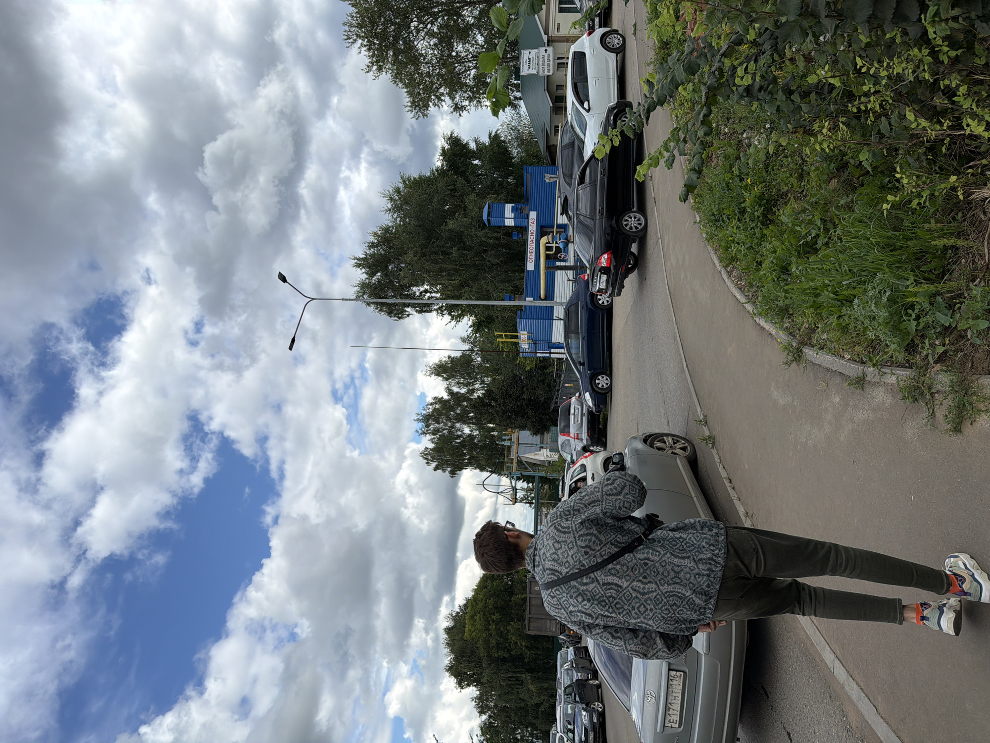 This is a full-shot, eye-level, outdoor photograph taken during the daytime in Yelabuga, Russia. In the foreground, a young man with short, dark hair and wearing glasses walks away from the camera, his back to the viewer. He is dressed in a patterned, long-sleeved shirt, dark green pants, and multi-colored sneakers. A dark bag is slung across his right shoulder. The man is walking on a paved sidewalk adjacent to a street with several parked and moving vehicles.
The street features a variety of cars, including a silver sedan in the foreground on the left, a black sedan, and a white hatchback on the right. Beyond the street, there are trees and buildings. A blue industrial structure with yellow pipes and white lettering that reads "ОГНЕОПАСНО-ГАЗ" (Flammable Gas) is visible, along with other smaller buildings and a street lamp. The sky is partly cloudy with a mix of white and gray clouds against a blue sky. The overall environment appears to be an urban or semi-urban setting with a functional industrial element.