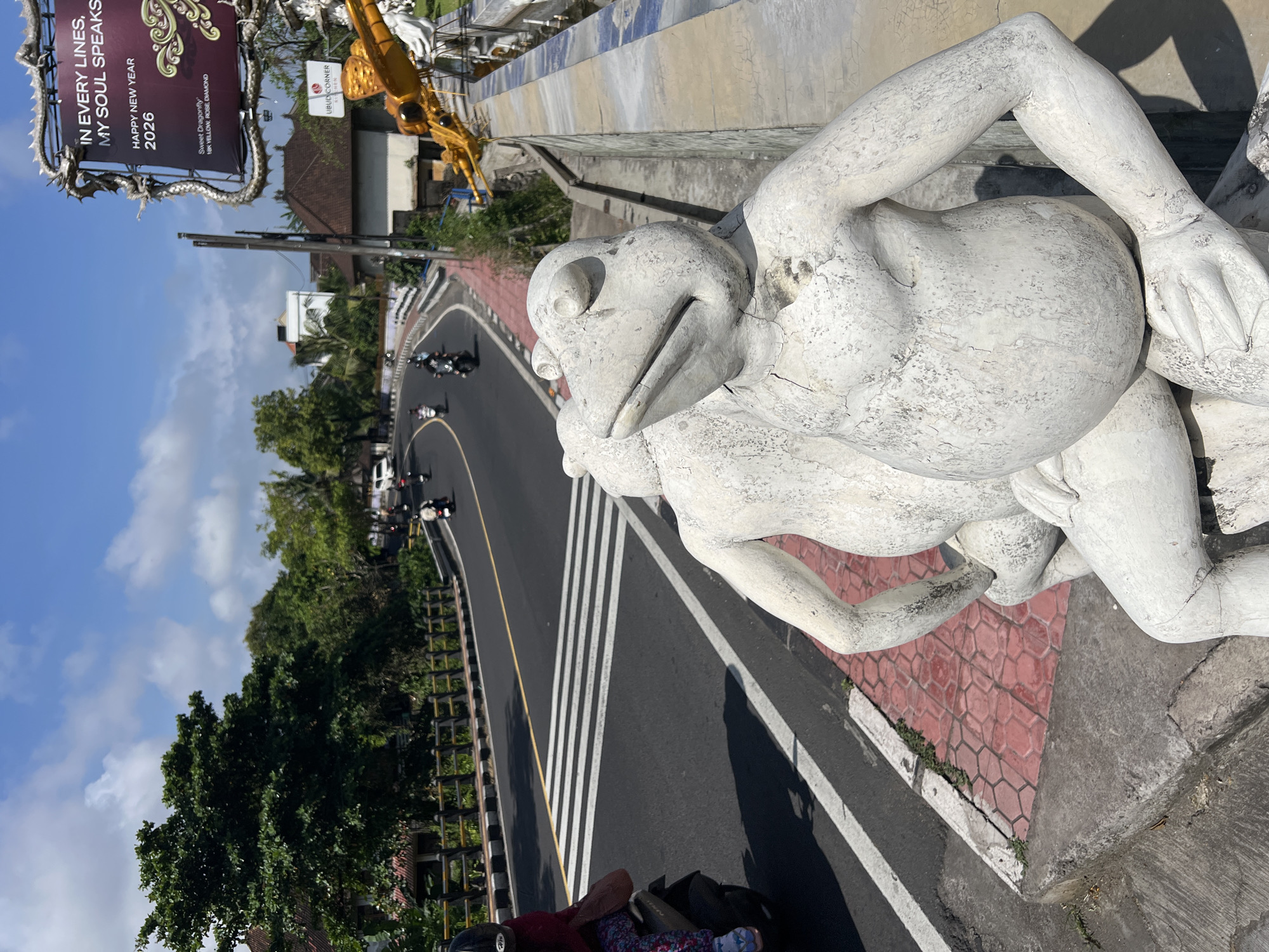 The image, taken in Ubud, Indonesia, shows a concrete frog statue in the foreground, positioned on a roadside platform. The statue is weathered and cracked. In the background, a road curves through a green, tropical landscape. Motorbikes and cars travel along the road, marked with white stripes and a yellow dividing line. A black and silver billboard stands to the left, advertising "Ubud Corner" and wishing "Happy New Year 2026." The sky above is partly cloudy.