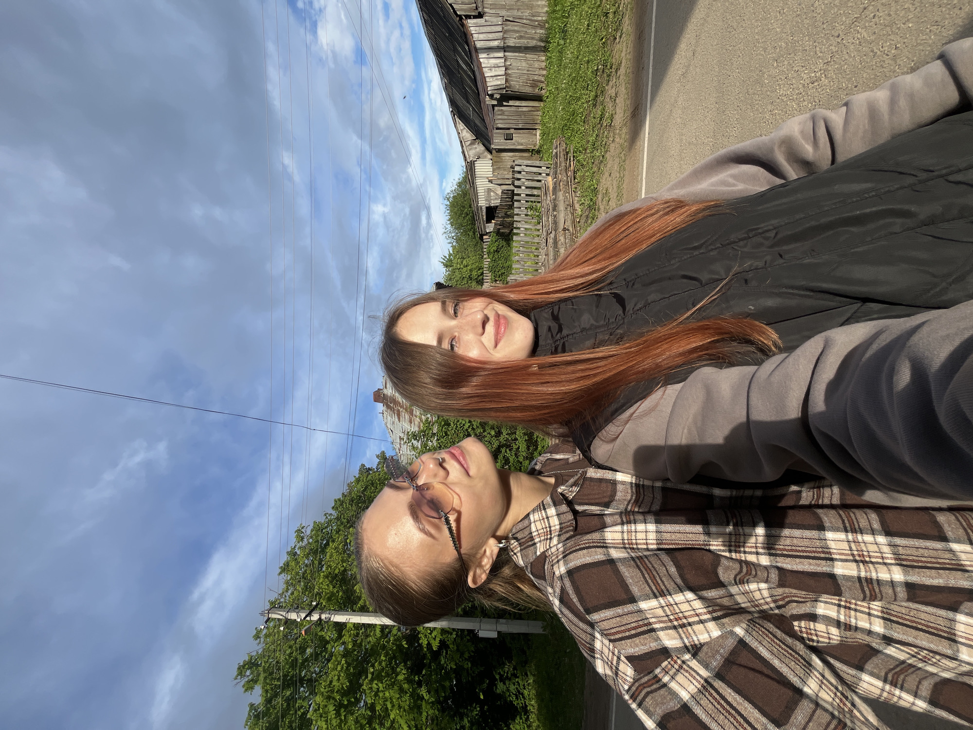 This image features two young women captured outdoors in what appears to be a rural or semi-rural setting in Foki, Russia. The scene is illuminated by daylight under a partly cloudy sky, with patches of blue visible.
In the foreground, one woman is seen in profile, looking slightly upwards with a relaxed expression. She has light brown hair, pulled back, and wears a brown and beige plaid shirt with glasses. A small nose piercing is also visible. Beside and slightly behind her, the second woman smiles warmly towards the camera. She has long, reddish-brown hair and is dressed in a black puffer vest over a grey long-sleeved top.
The background elements suggest a traditional village or countryside environment. To the right, a weathered wooden fence borders a dirt road or path. Beyond the fence, rustic wooden buildings, possibly old houses or barns, stand amidst green trees and grass. Overhead, multiple power lines crisscross the sky, characteristic of rural infrastructure. The overall atmosphere conveys a quiet, sunlit day.
No visible text is present in the image.