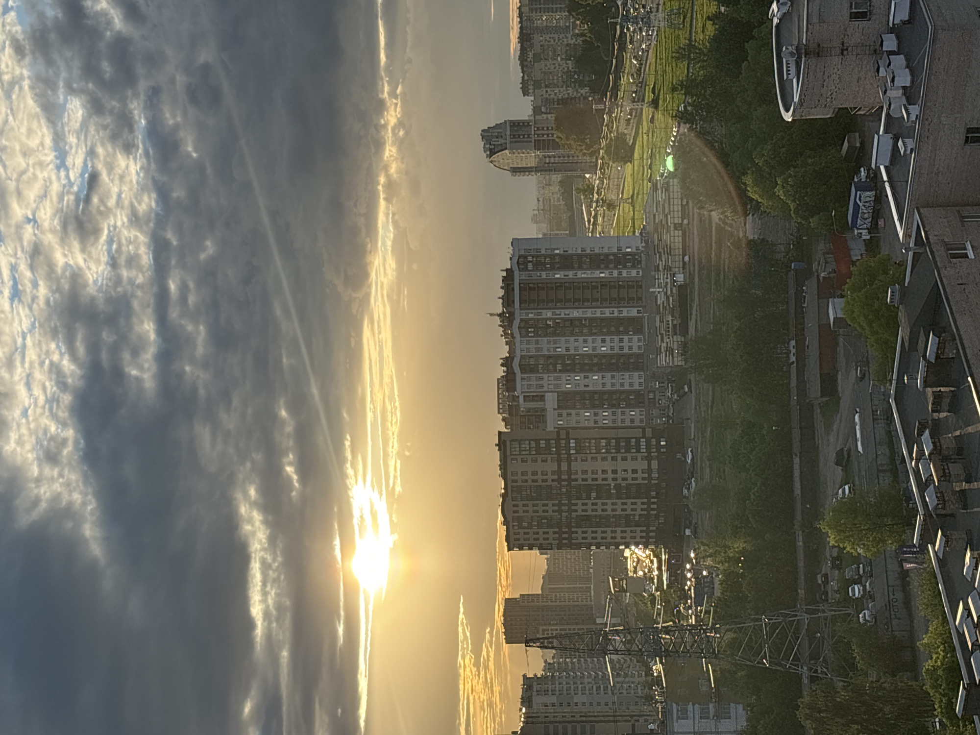 The image depicts an urban landscape at sunset. The sun is positioned low in the sky, casting a golden glow across the scene. A mix of dark, heavy clouds and bright, sunlit clouds dominate the left side of the image. On the right side, there are several high-rise buildings, some appearing residential. A park with trees and paths is visible in the distance behind the buildings. Utility poles and power lines are situated in front of the buildings. The location appears to be in a city, but the specific city and country are unknown.