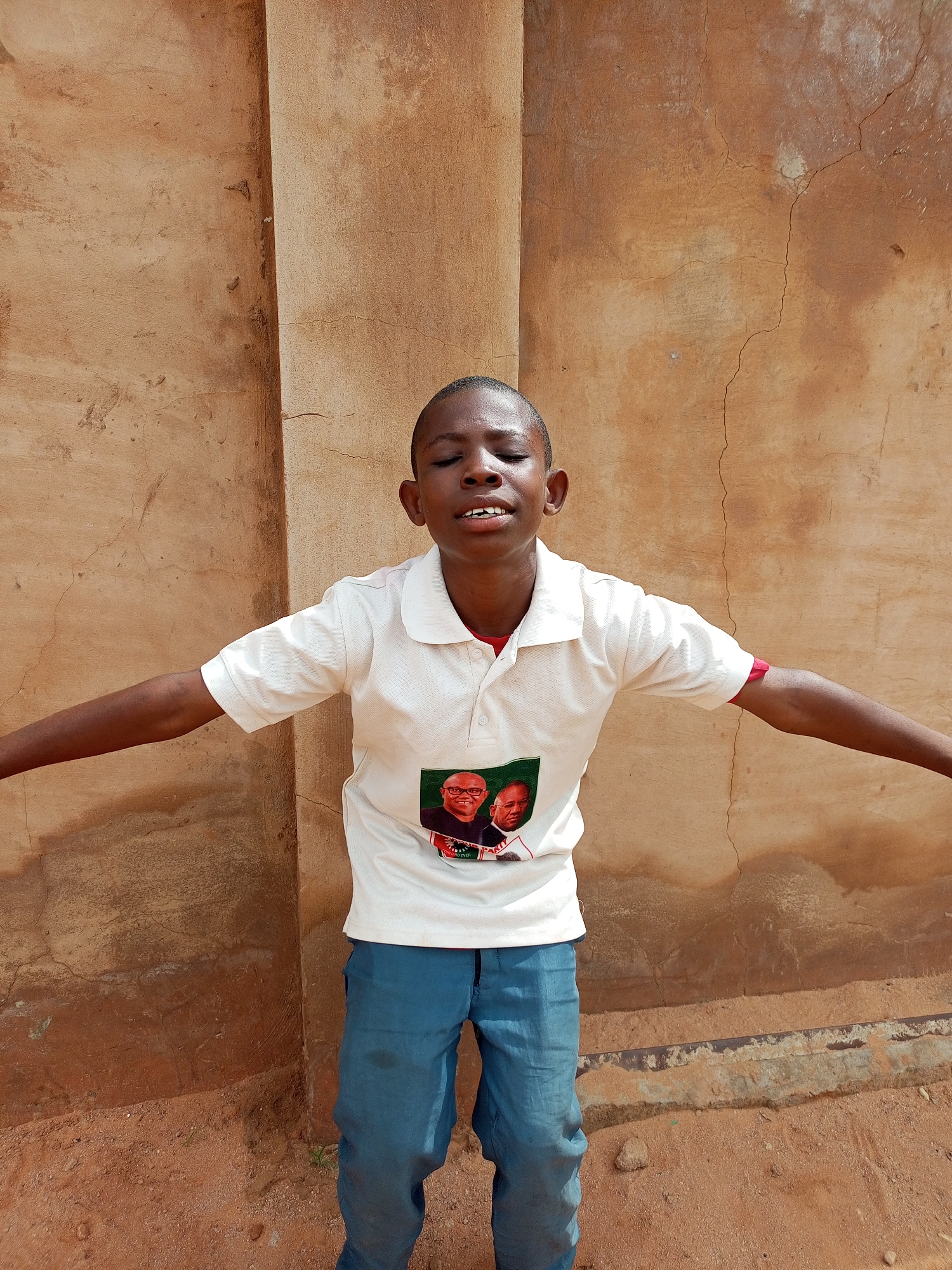 A young boy stands with his arms outstretched and eyes closed in front of a textured, light brown wall with visible cracks. He wears a white polo shirt with a red collar and a small image of two men on the left side of his chest. The boy has short, dark hair and is wearing blue trousers. The ground in front of the wall is composed of dirt and small stones. This scene is located in Tempure, Nigeria.