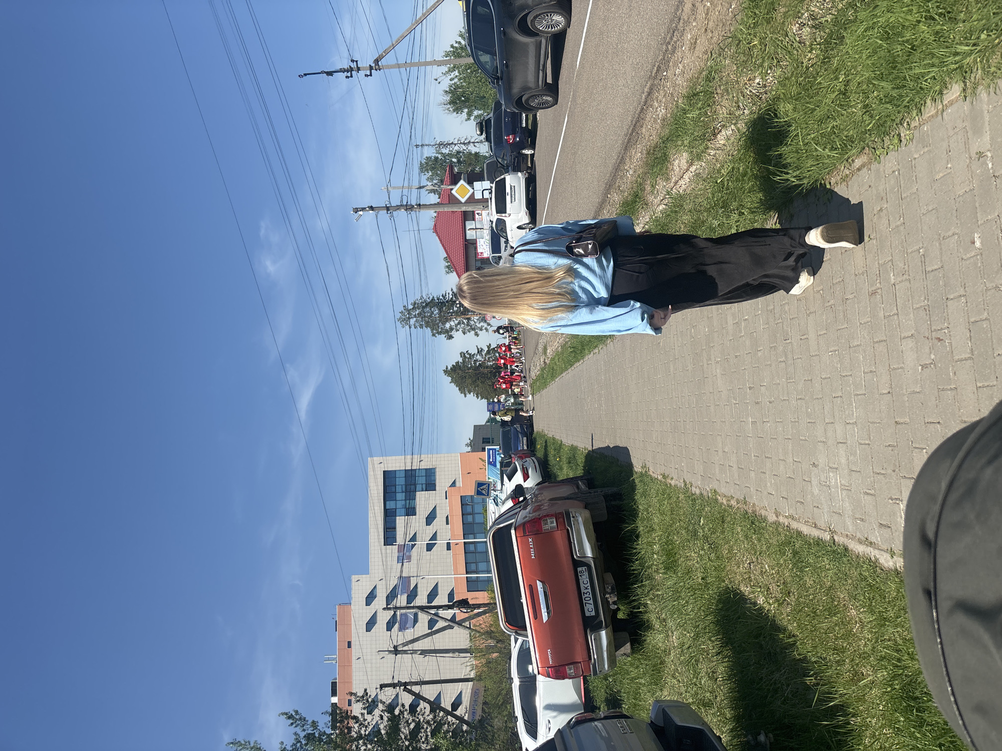 The image shows a blonde woman walking on a brick sidewalk in Прикамский, Russia. She is wearing a light blue shirt and a black skirt. A row of parked cars lines the street to her right. Buildings and power lines are visible in the background, under a clear blue sky. The grass separates the road from the sidewalk.