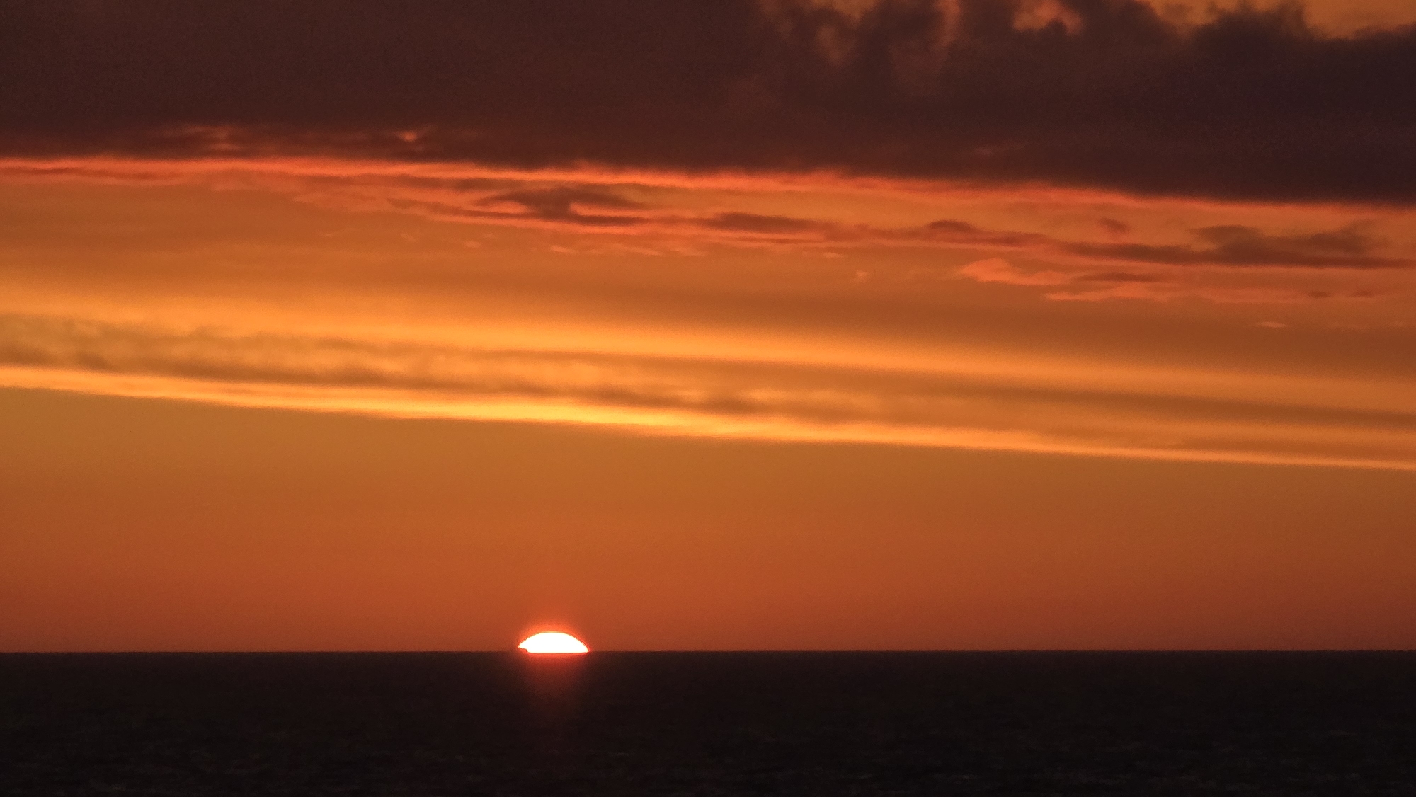 The image captures a sunset over the ocean. The sun, a semi-circular orange orb, is positioned just above the horizon line. The sky transitions from deep red and orange hues near the top to a lighter orange closer to the horizon. Clouds are present in the upper portion of the image, varying from dark shades to brighter, more translucent bands of orange. The ocean is a dark, nearly black, band at the bottom of the frame. No people, specific objects, or actions are visible beyond the natural scene of a sunset. The location and city are unknown.