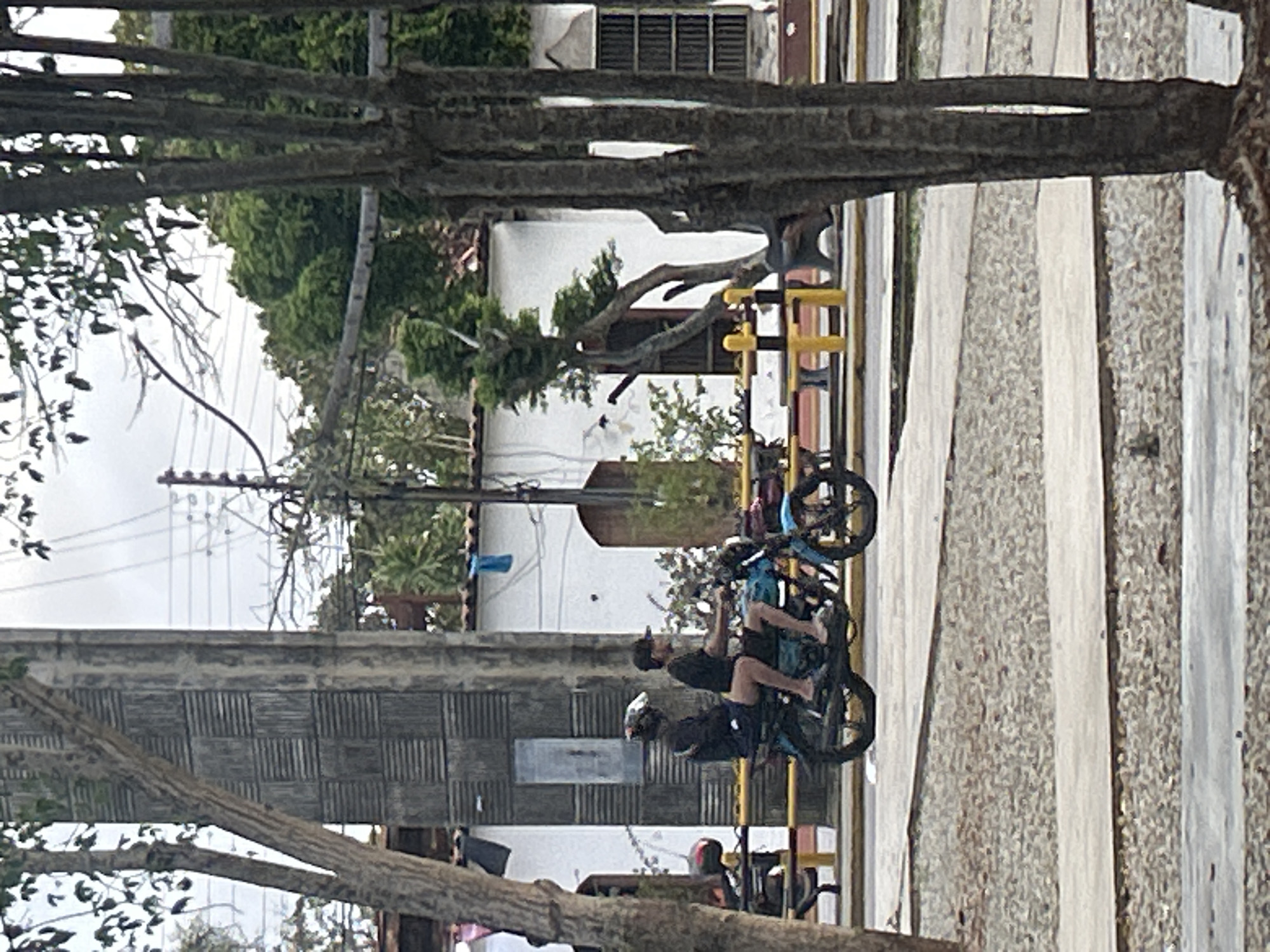 Two individuals are riding on a blue motorcycle on a street in Santa Ana, Venezuela. The person in the front is wearing a dark shirt and a black baseball cap, while the person in the back is wearing a dark shirt and a helmet. They are both wearing shorts and flip-flops. The motorcycle is moving from left to right. In the background, there is a white building with arched doorways and windows. There are also trees and utility poles. The street appears to be paved with asphalt, and there is a gravel area to the left of the street. A yellow barrier with black posts is visible along the side of the street.