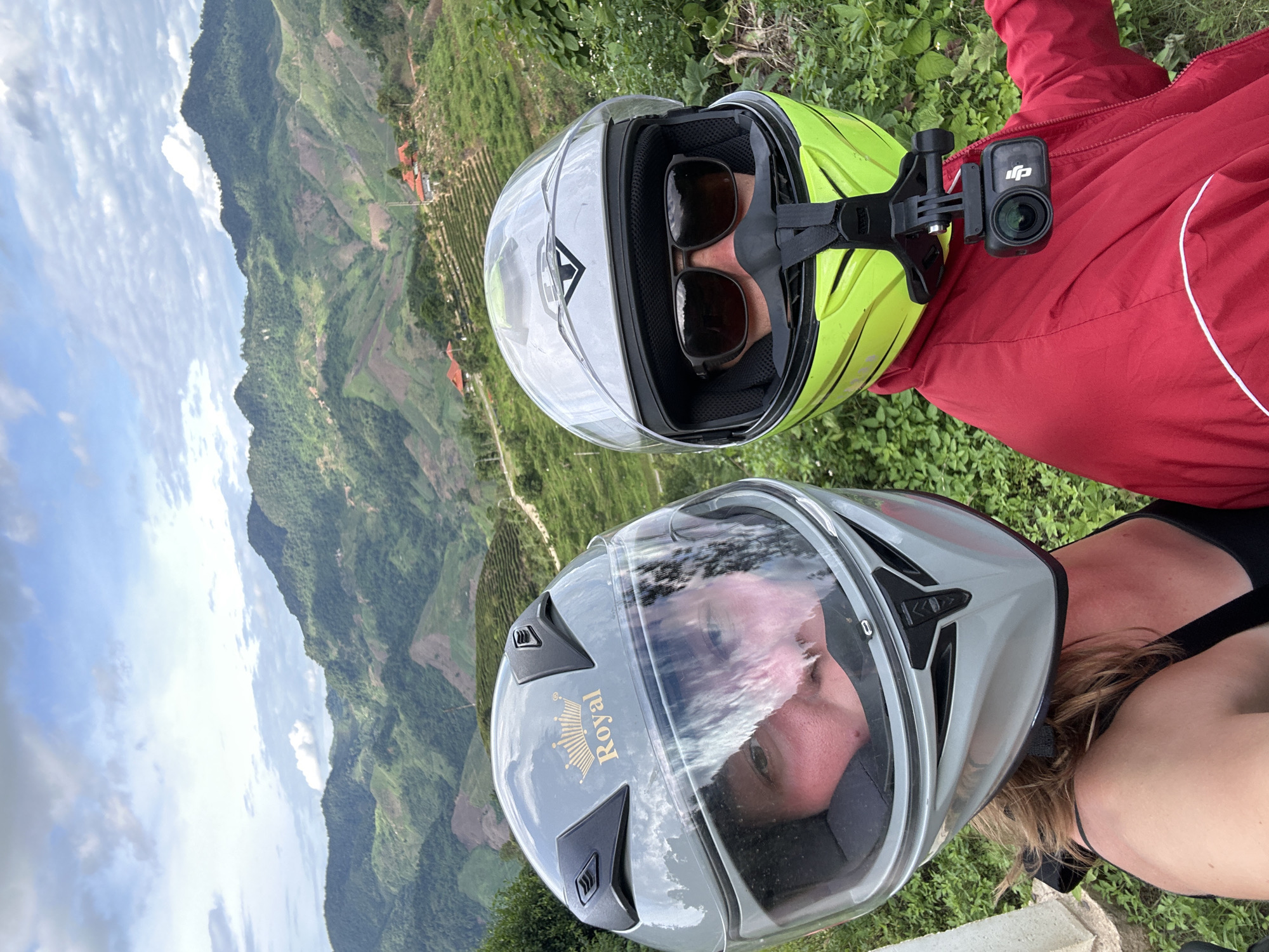 Two individuals wearing motorcycle helmets pose for a selfie with a scenic mountain landscape in the background, identifiable as the terraced hills of To Mua, Vietnam. The person on the left wears a gray helmet with a "Royal" emblem and their face is partially visible through the visor, showing their eyes and flushed cheeks. They are wearing a black tank top. The person on the right wears a bright yellow-green helmet with a white emblem and dark sunglasses. A small action camera, a DJI Osmo Action, is mounted on their helmet. They are wearing a red jacket. The background reveals lush, green, terraced hillsides with some scattered houses. The sky above is partly cloudy with a mix of blue sky and white clouds. The image captures a moment of adventure and sightseeing in a mountainous region.