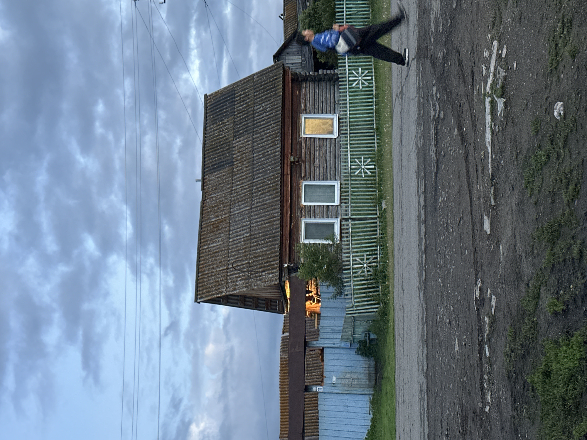In an outdoor, overcast scene in a rural setting, a wooden house with a corrugated roof stands behind a green and white picket fence. Three windows are visible on the house; one is dark, one reflects the sky, and the third glows with interior light. A person walks from right to left in front of the fence, wearing a blue hooded jacket and dark pants, carrying a bag over their shoulder. The ground in front of the fence is paved and appears to be a dirt road with some puddles. A blue wooden gate is visible to the left of the house, with a visible number "11" on it. Power lines are strung across the sky.