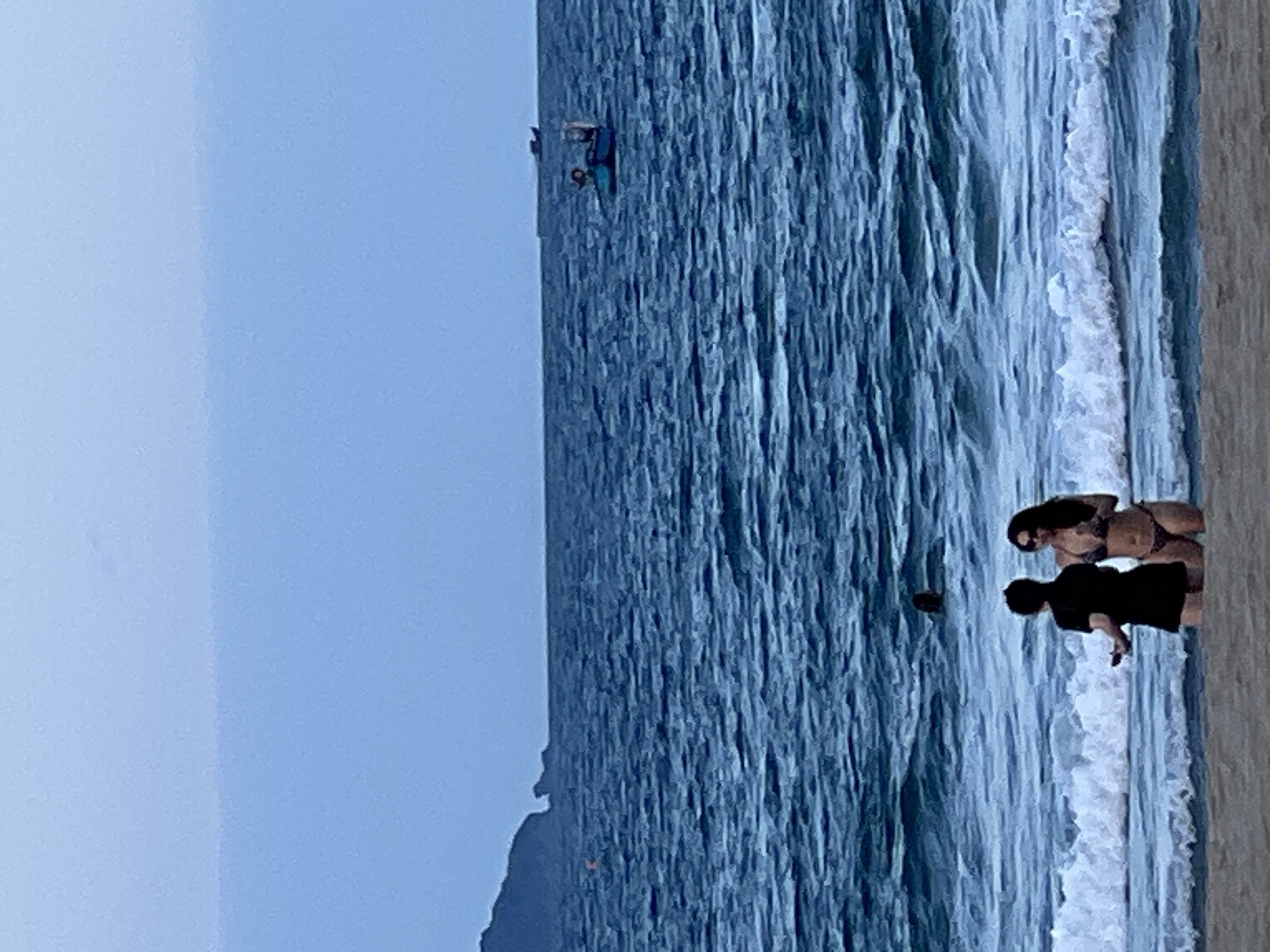 The image depicts a serene beach scene with a gentle sea and a hazy sky, characteristic of Đà Nẵng, Vietnam. In the foreground, two individuals, one wearing a black t-shirt and shorts, and another in a bikini, stand near the water's edge, appearing to engage in conversation. The water is a deep blue with moderate waves lapping at the shore. Further out at sea, a small boat with people on it is visible, along with another distant boat. A dark, mountainous silhouette of land juts out from the left side of the frame, suggesting a coastal landscape typical of Đà Nẵng's environs. The overall atmosphere is tranquil, with the soft light indicating either early morning or late afternoon.