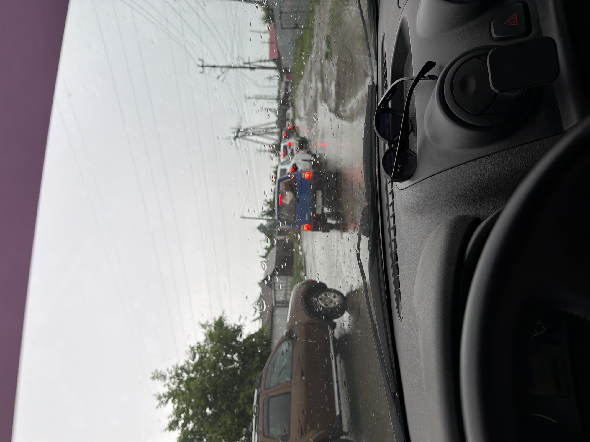 The image is taken from inside a vehicle looking out through a rain-streaked windshield. The perspective is from the driver's seat, with the steering wheel and dashboard visible in the foreground. Sunglasses rest on the dashboard.
Outside the vehicle, a brown SUV is in the left lane, with its front end cut off by the frame. The road is wet and reflects the overcast sky, suggesting it is raining or has recently rained. Traffic is backed up, with several cars visible in the distance, their brake lights illuminated. A blue car is prominently featured in the middle lane, ahead of other vehicles.
The environment appears to be a residential area, with houses and fences visible along the right side of the road. Utility poles with power lines are also present. The sky is a uniform gray, consistent with rainy weather.
While the specific location within the city of Chaikovsky, Russia, cannot be determined from the image alone, the scene depicts a common urban or suburban environment experiencing inclement weather and traffic congestion. No individuals are clearly visible within or outside the vehicles.