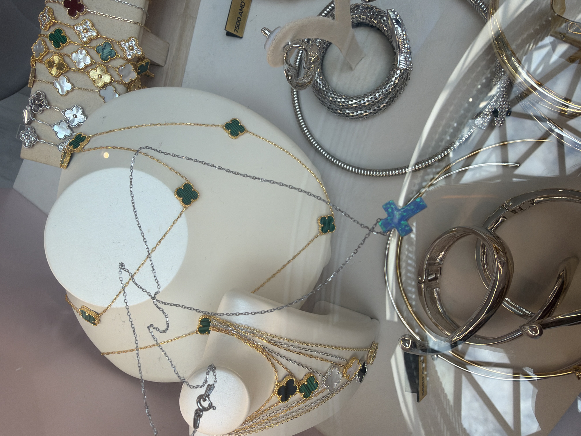 The image shows a display of jewelry, likely in a shop window. There are necklaces with clover-shaped pendants, a silver snake-like bracelet, and various silver bangles. One of the price tags shows "7500 AMD", which is the Armenian Dram currency, suggesting that the location is likely in Yerevan, Armenia. The jewelry is displayed on a white mannequin bust and stands. The scene is brightly lit, with reflections on the glass.