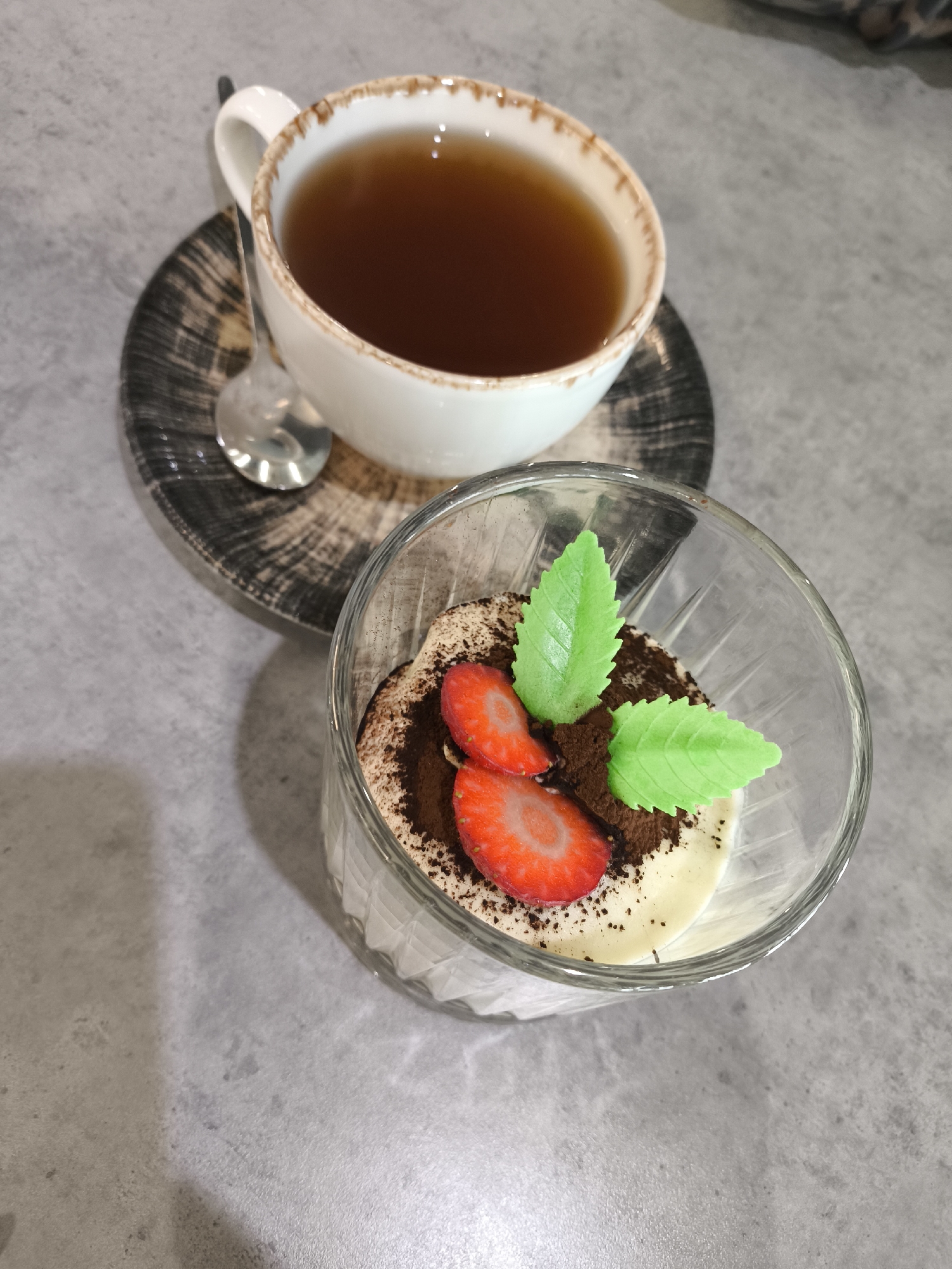 The image shows a teacup on a saucer with a spoon, and a glass dessert dish on a grey surface. The teacup is white with a brown rim and contains a dark liquid, presumably tea. The saucer is patterned with black and brown. A silver spoon rests on the saucer next to the teacup. The dessert dish contains a white cream-like base, topped with cocoa powder and two slices of strawberry and two artificial mint leaves. The background is a solid grey surface. The image was taken indoors. There is no indication that the image was taken in Izhevsk, Russia.