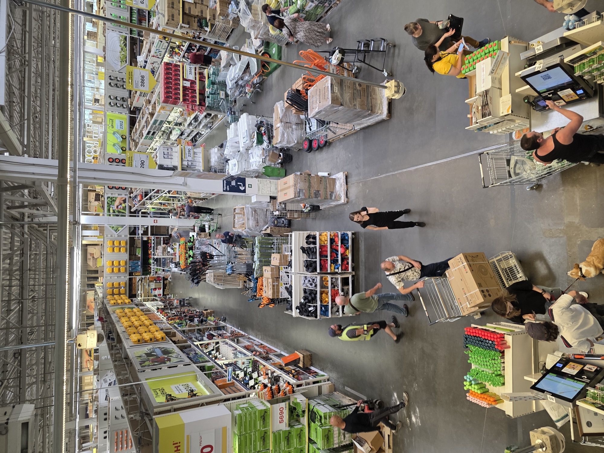 An overhead, wide-angle shot captures a brightly lit, large retail store's interior with rows of merchandise and shoppers. The store appears to be a hardware or home improvement store, with aisles stocked with items like tools, gardening equipment, cleaning supplies, and building materials. In the foreground, two shoppers are at a checkout counter, one of whom is interacting with a payment terminal. A small dog on a leash stands patiently beside them. In the midground, several shoppers are pushing shopping carts or walking through the aisles, some examining products. The background shows more shelves and displays, extending deep into the store. The overall environment is clean and organized, with a high ceiling and industrial-style lighting.
