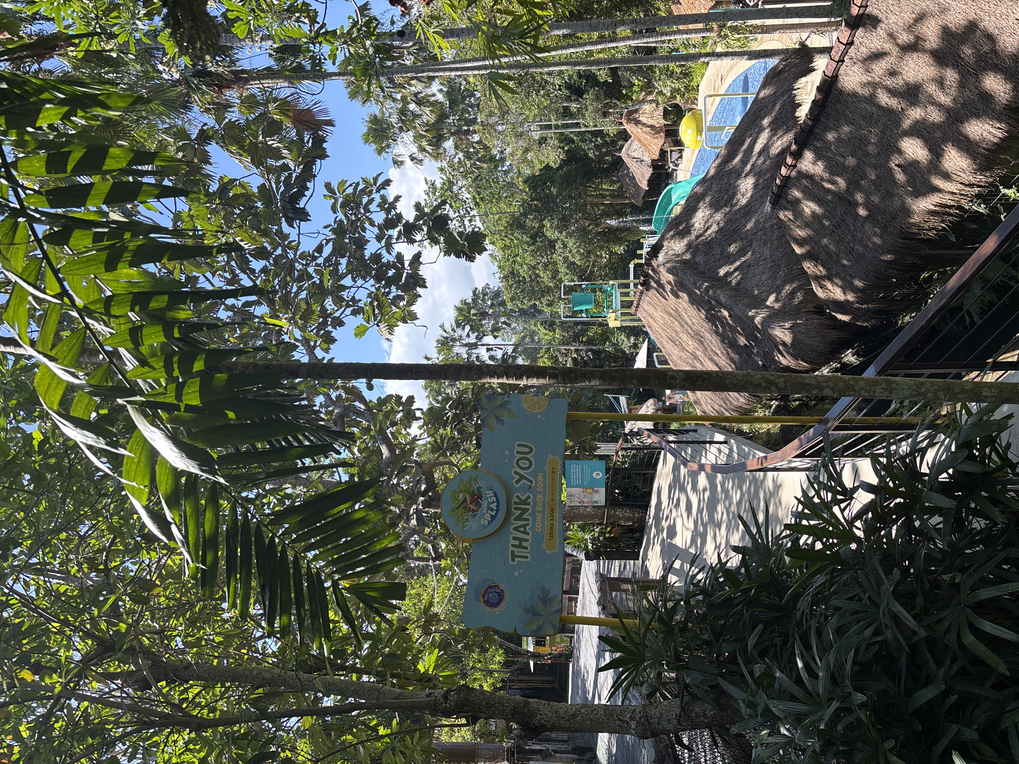 The image shows a sign with "SPLASH" written on it, along with "THANK YOU" and "COME BACK SOON". Below that, there is text in Indonesian: "TERIMA KASIH!" and Japanese: "ありがとうございます". The sign is light blue with green and yellow accents and is supported by yellow poles. It is situated in an outdoor environment with lush green trees and palm trees. In the background, there is a glimpse of a water park with a blue pool, a water slide, and thatched-roof huts. The overall scene suggests a tropical location, possibly a tourist attraction. The sign and the water park elements strongly suggest that this image was taken at the Bali Splash Water Park in Sukawati, Indonesia.