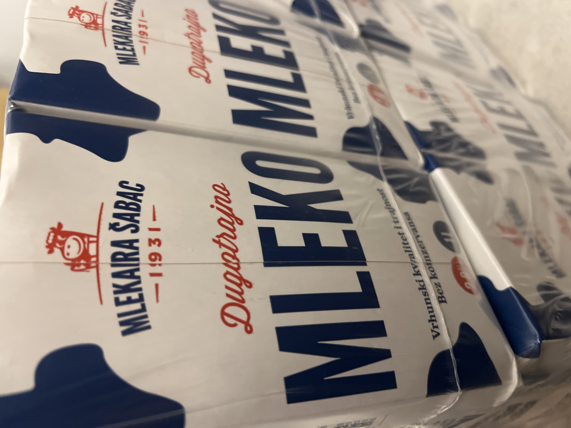 The image shows a close-up of stacked milk cartons, likely in a store or warehouse setting. The cartons are white with blue and red lettering. Prominently displayed on the cartons is the text "MLEKARA ŠABAC" and "DUGOTRAJNO MLEKO" in large, bold, blue lettering. The brand appears to be from Šabac, Serbia, indicated by "ŠABAC" in the text. There is also a stylized red cow logo on some of the cartons. The cartons are wrapped in clear plastic. The lighting suggests an indoor environment. There are no people visible in the image. The image is a static shot focused on the product packaging. The location of Budva, Montenegro is not visually represented in the image itself.