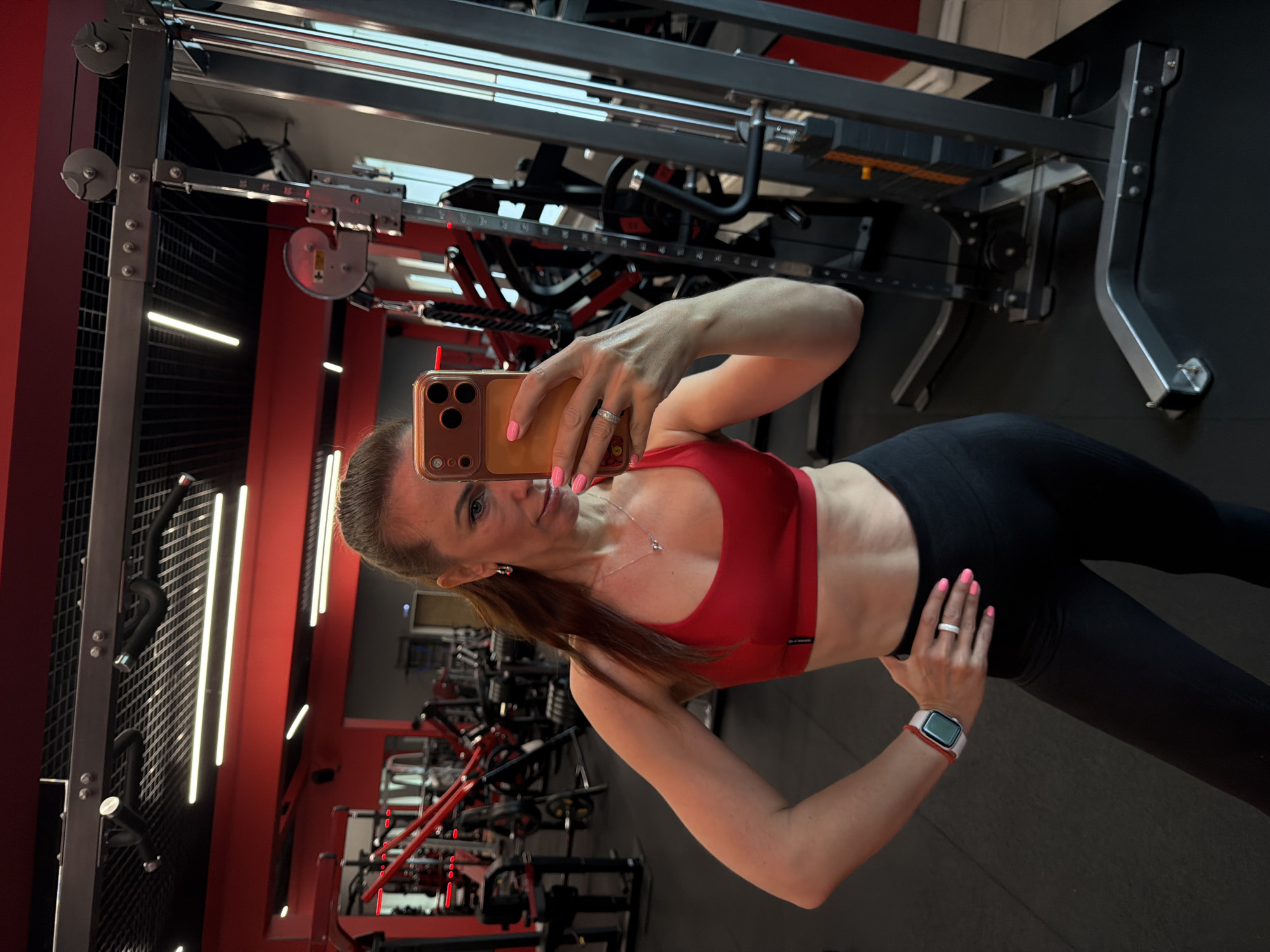 A woman with brown hair tied in a ponytail, wearing a red sports bra and black leggings, stands in a gym and takes a selfie in a mirror. She is wearing a smartwatch on her left wrist and a ring on her right ring finger. The gym has red walls and black equipment, including weight machines and pull-up bars. The floor is black rubber matting. The lighting appears to be from fluorescent ceiling lights and the machines. The image is taken from a medium shot, capturing the woman from the waist up and showing some of the gym equipment in the background.