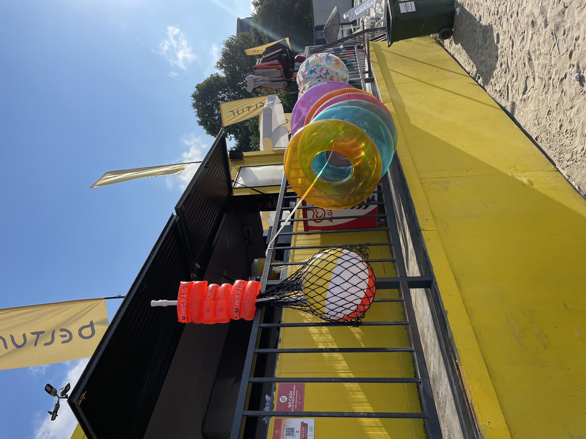 The image depicts a beachside kiosk on a sunny day in Kadıköy, Turkey. The kiosk is painted bright yellow and has a black overhang. Several inflatable swimming rings in various translucent colors, including orange, yellow, purple, and aqua, are hanging from a railing in front of the kiosk. To the left, a net bag contains a red and white beach ball. Above the beach ball, a stack of red inflatable rings is suspended. Two yellow flags with black text, possibly advertising, are visible. One flag hangs from the kiosk, and another is further back. The environment includes a sandy beach in the foreground and trees in the background. A green trash can is positioned to the right of the kiosk. The overall scene suggests a recreational area, likely a beach or pool, equipped for summer activities.