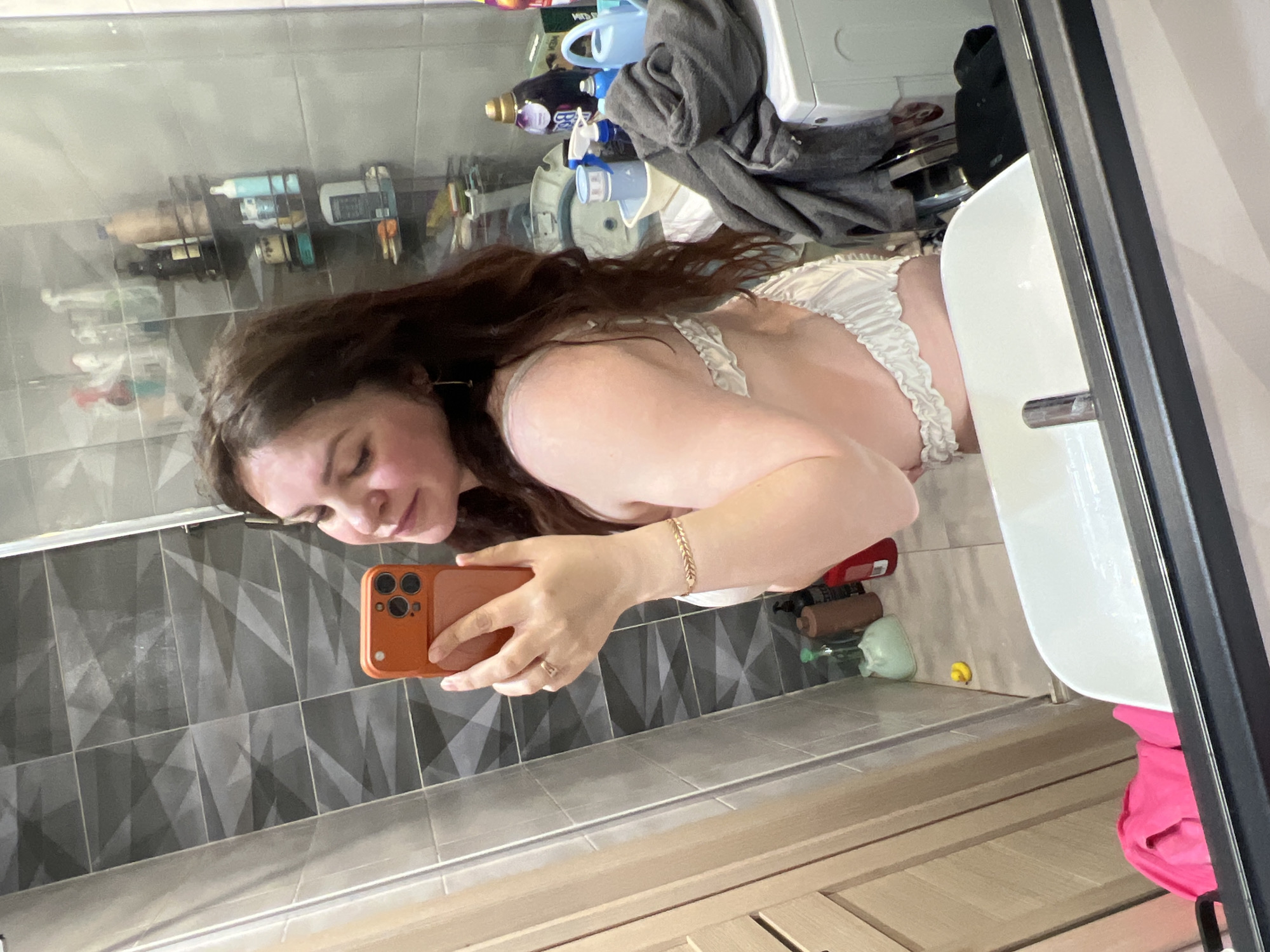 A reflection in a bathroom mirror shows a woman holding an orange smartphone, presumably taking a selfie. She is wearing white lingerie. The bathroom has gray tiled walls, a white sink, and a glass shower door. In the background, shampoo bottles are visible on a shelf. The city of Noviy, Russia is not confirmed as the location, as there is no definitive evidence to support it.