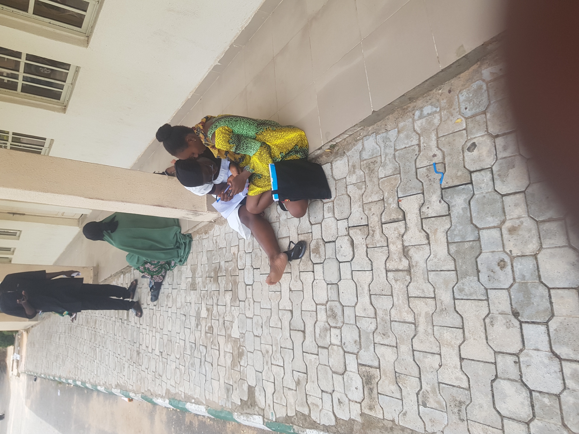 The image depicts an outdoor scene in Dabawa, Nigeria. A woman in a yellow and green patterned dress is kneeling, leaning over and looking at a person wearing a white shirt and face mask who is sitting on the ground. They appear to be reviewing documents. A black bag is near them.
In the background, two other people are visible, standing on the walkway. One is dressed in a black suit, and the other is wearing a green garment.
The setting appears to be a paved walkway adjacent to a light-colored building with windows. The ground is paved with interlocking stones.