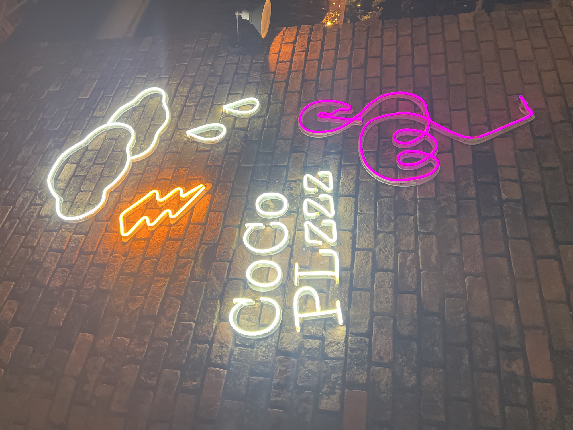 This media file showcases a neon sign displayed on a brick wall at night. The sign features various illuminated elements, including two white cloud-like shapes, three white teardrop shapes, and an orange lightning bolt shape. Below these, a vertical arrangement of white neon letters spells out "COCO PIZZZ". To the right of the text, a bright pink neon shape forms a stylized swirl or musical note. The brick wall suggests an outdoor setting, possibly a restaurant or bar, with a spotlight casting an orange glow on the top left portion of the wall. The overall scene is dark, indicating nighttime, and the neon lights provide the primary illumination. There are no people visible in the frame. The context of Baan Bo Phut, Thailand, suggests a tropical or coastal environment, though the image itself does not contain explicit visual cues for this. The combination of neon signs and brickwork creates a vibrant and somewhat playful atmosphere.