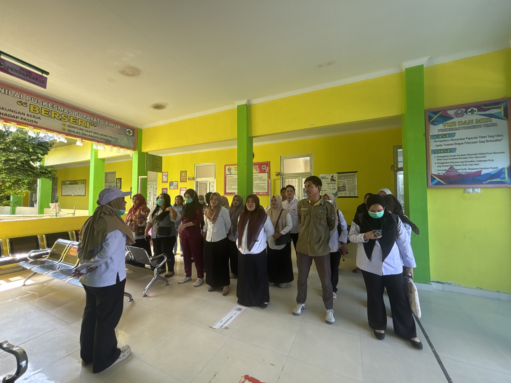 The image depicts a group of people, predominantly women, standing in a well-lit indoor area that appears to be a waiting room or common area of a medical facility. The environment is characterized by bright yellow and green walls, white tiled flooring, and a high ceiling with exposed beams and lighting fixtures. Several individuals are wearing masks, suggesting a health-conscious setting.
In the foreground, a person wearing a light blue long-sleeved shirt, black pants, and a light brown headscarf is facing away from the camera, with their hands clasped behind their back. To their left, a row of metallic waiting chairs is visible.
Behind the foreground individual, a diverse group of people are standing in a line or cluster, some are looking towards the camera, while others are engaged in conversation or looking in different directions. Many of the women are wearing headscarves and are dressed in professional attire. One woman in the center wears a dark brown headscarf and a white shirt with a black skirt. Another woman on the far right wears a maroon outfit and a light brown headscarf.
Various posters and signs are visible on the walls. On the left side of the image, a banner with text in Indonesian reads "NILAI PUSKESMAS POPAYATO TIMUR" and "BERSERI". Another sign above it indicates "RUANG PENDAFTARAN". On the right wall, a prominent poster titled "VISI DAN MISI PUSKESMAS POPAYATO TIMUR" is displayed, outlining the vision and mission of the facility. Other smaller signs and posters with Indonesian text are also present.
The overall impression is a gathering of staff or patients in a clean and organized public health service building. The location is identified as Telaga, Indonesia, likely within the Puskesmas Popayato Timur facility. The atmosphere appears to be calm and orderly.