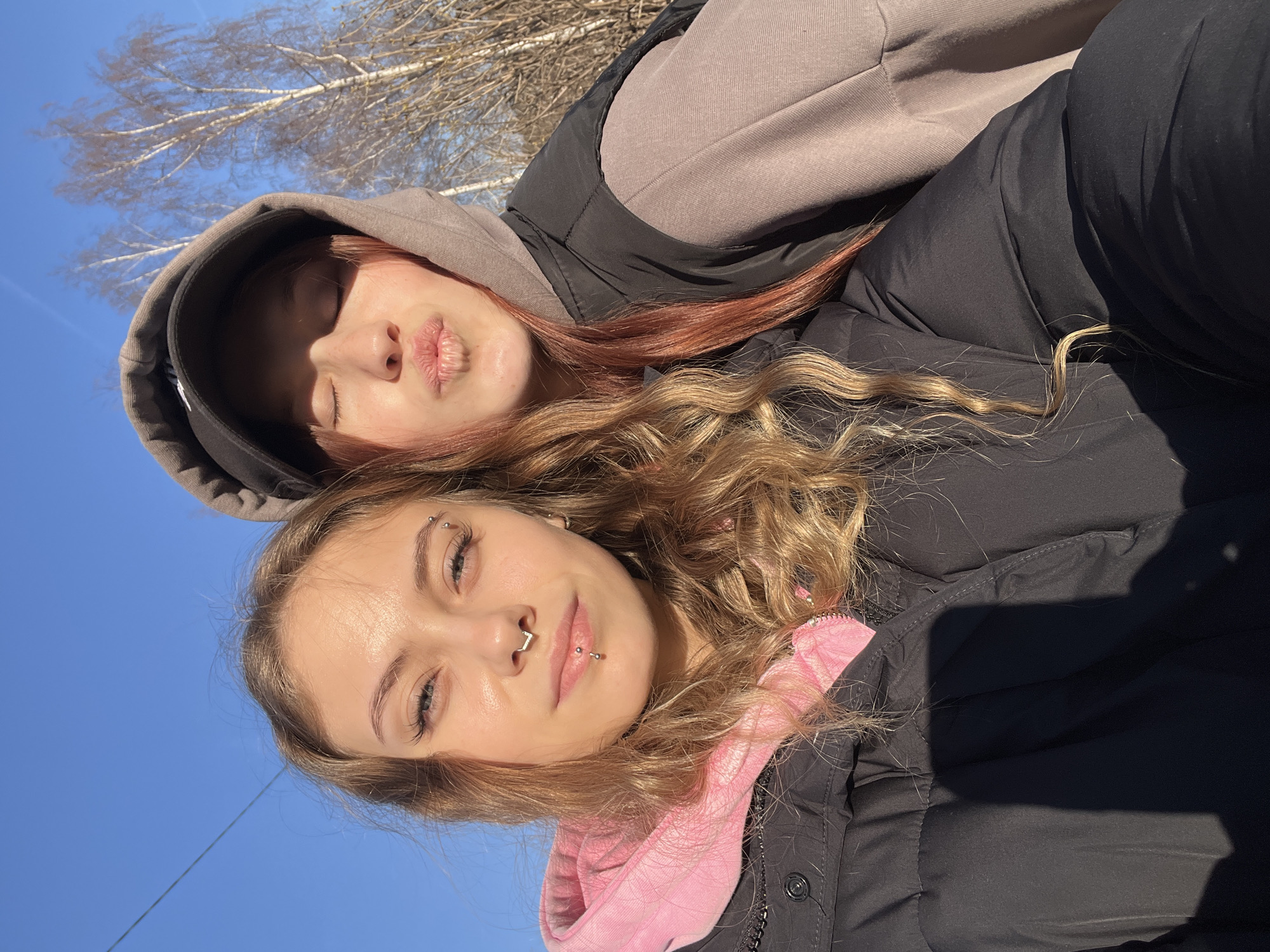 Two young women are posing for a selfie outdoors, likely in Фоки, Russia given the prompt. The foreground woman has blonde, curly hair and visible facial piercings on her eyebrow, nose, and lip. She is wearing a black jacket with a pink hooded sweatshirt underneath. The background woman has red hair and is wearing a grey hooded sweatshirt and a black jacket. The sky is blue and there are bare trees visible in the background.