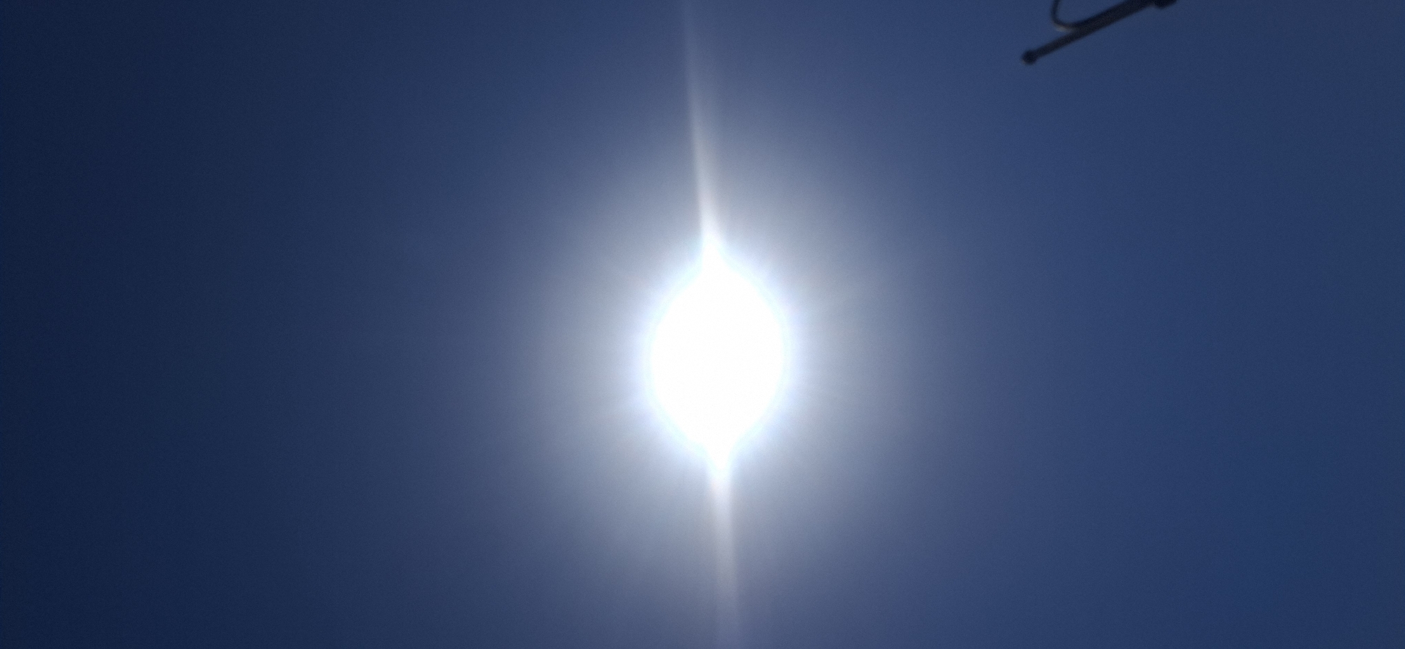 A bright, overexposed sun is centered in the frame, creating a lens flare effect that extends horizontally across the dark blue sky. At the bottom right corner of the image, a portion of a dark, thin pole-like object is visible, angled upwards. The scene appears to be an outdoor environment, possibly during daylight hours given the sun's prominence. No specific location in Yerocham, Israel is evident from the image content.
