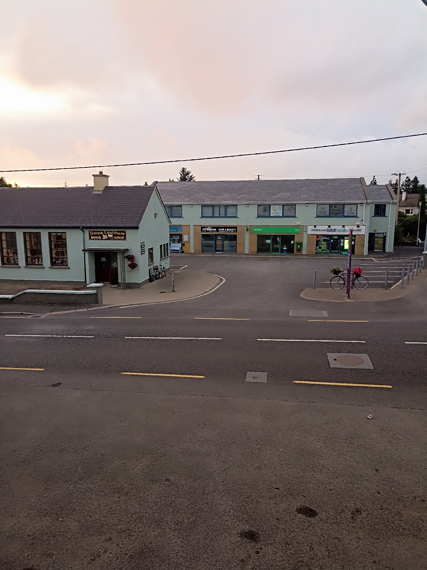 The image depicts a street view in Annagry, Ireland, likely during overcast conditions. In the foreground, a paved road features yellow and white lane markings. Further back, a small shopping complex is visible, with several businesses housed in a light blue building. One storefront clearly displays a sign reading "SIOPA LEADHÁIR BOOK SHOP". Adjacent to this is a series of other businesses, including "ALLURE HAIR & BEAUTY", "an post", "CO.GARLAINN", and "TOTALHEALTH PHARMACY". Parked in front of the shopping complex is a bicycle adorned with flower baskets. The sky above is a pale, washed-out color, suggesting either dawn, dusk, or heavy cloud cover. Trees and a house are visible in the background to the right.