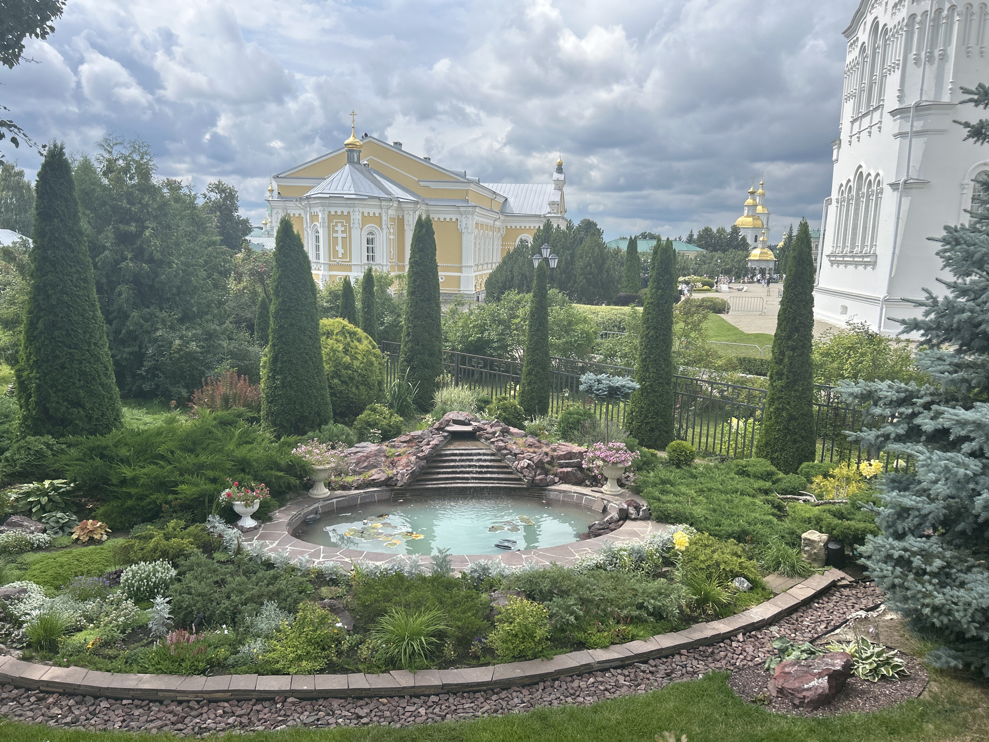 The image depicts a meticulously landscaped garden with a pond and waterfall feature in the foreground. Surrounding the pond are various types of evergreen shrubs, low-lying ground cover plants, and ornamental grasses. Two white decorative urns with pink flowers sit on either side of the waterfall. In the background, a large, pale yellow church with a golden dome is visible, along with other architectural elements of what appears to be a religious complex. The sky is overcast with dramatic clouds. The scene is likely part of the Holy Trinity Seraphim-Diveyevo Monastery in Diveyevo, Russia, a significant pilgrimage site. There are no people clearly visible in the image, and the environment suggests a serene and well-maintained spiritual setting.