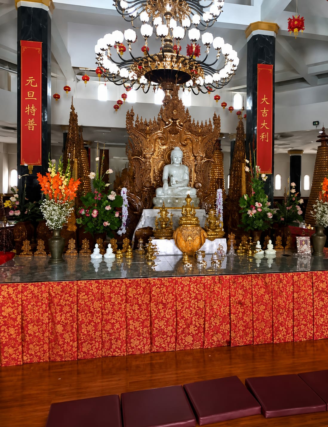 The image depicts the interior of a Buddhist temple, specifically in the city of Dolat Rayat, Indonesia. The central focus is a white Buddha statue seated on a golden, ornate throne. The throne is highly detailed with intricate carvings and is flanked by smaller golden statues and ornate spires. In front of the throne, a large, decorative golden urn or vessel is placed on a marble surface. The altar area is adorned with various offerings, including flowers in vases, and smaller statues of Buddha. Red banners with gold lettering hang from tall, dark pillars on either side of the altar. A large, ornate chandelier hangs from the ceiling, illuminating the scene. Red lanterns are also visible, suspended from the ceiling and along the upper walls. The altar is covered with a red cloth featuring a gold floral pattern. In the foreground, several dark red floor cushions are arranged on a polished wooden floor. The overall impression is one of reverence and ornate religious decoration. There are no people visible in the image.