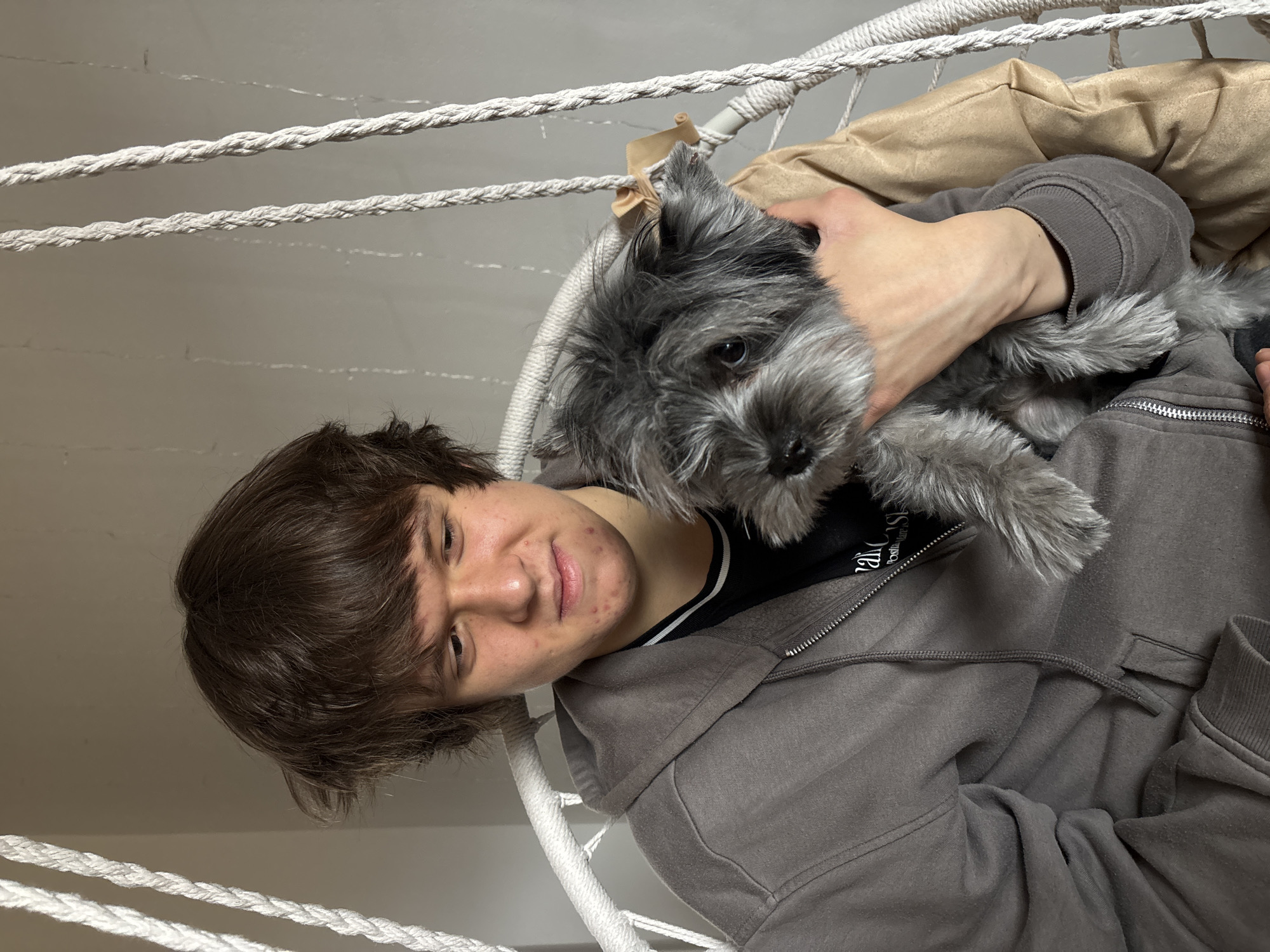 A young man is pictured with a small dog, both seated in what appears to be a hanging swing chair or hammock, indicated by prominent white braided ropes in the foreground and background. The image is rotated, showing the man leaning slightly to his left and the dog held in his arms. The setting is indoors, with a light gray or off-white wall visible in the background, partially obscured by a beige pillow or blanket positioned behind the man's head.
The man has short, dark brown hair and a light complexion, with some acne visible on his cheeks. He is wearing a grey hooded sweatshirt with a zipper down the front and a fuzzy grey lining around the hood opening. Underneath, a black t-shirt with a white stripe along the collar is visible. He has a gentle, closed-mouth smile and looks slightly to his right, towards the camera.
He is holding a small, fluffy dog with a mix of grey and black fur, dark eyes, and pointed ears. The dog is looking directly towards the viewer with a calm expression. Its fur texture suggests a breed like a miniature schnauzer or terrier mix. The man's right hand gently supports the dog's back, while his left arm cradles its body.
A small piece of light brown tape or fabric is tied around one of the white ropes near the dog's head. On the man's black t-shirt, partially obscured by the grey hoodie's zipper, faint white text is visible, reading "Original" and "POSI...", likely part of "Positive Case."
The scene captures a moment of companionship between the man and his pet, likely in a home setting in Chaikovsky, Russia.
