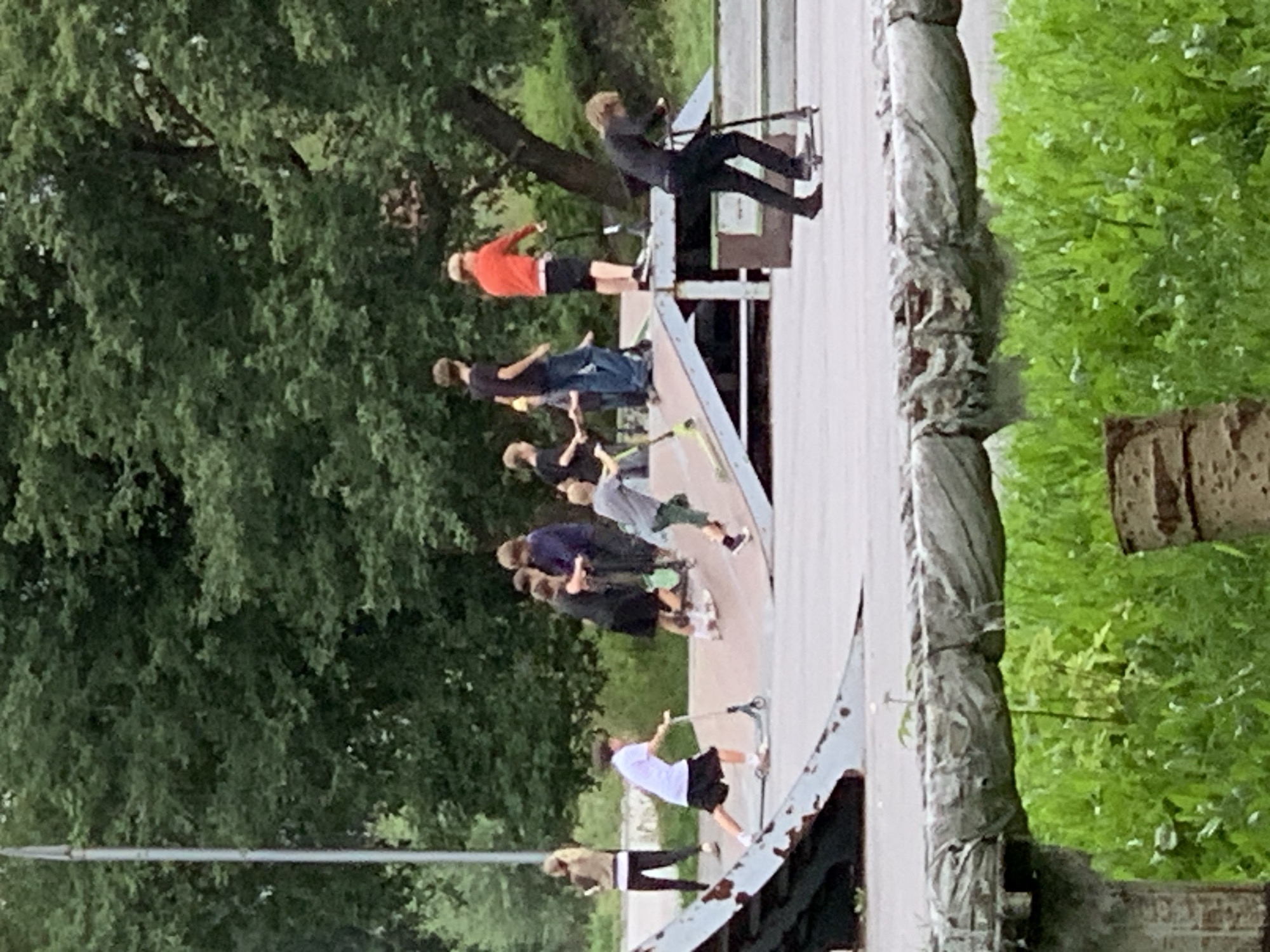 The image shows a group of teenagers at an outdoor skate park in Prokopyevsk, Russia, on an overcast day. Several individuals are riding scooters on ramps and flat surfaces. One person in the foreground is on a scooter, dressed in black. Another person in a white t-shirt and black shorts is also on a scooter, positioned on a ramp. A group of other teenagers are visible in the background, some on scooters and others standing. The skate park features concrete surfaces and metal ramps. Lush green trees and foliage surround the park, and a weathered, light-colored metal structure is visible in the lower foreground.