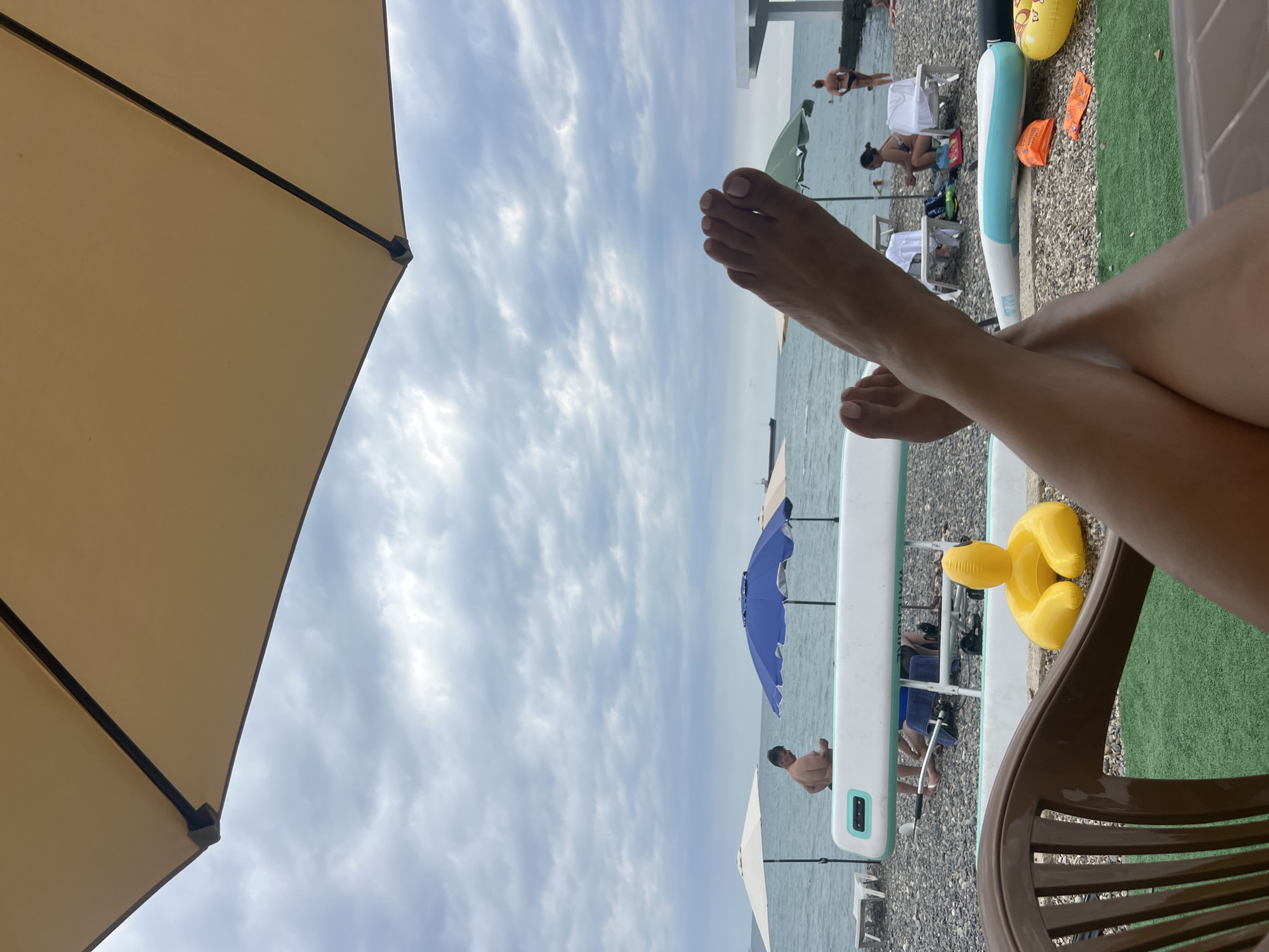 The image depicts a beach scene in Sokhumi, Abkhazia, under a cloudy sky. In the foreground, a person's legs and feet are crossed, suggesting they are relaxing in a chair. The camera angle is from a low perspective, looking out towards the sea. The beach is covered in pebbles, with patches of green artificial turf. Several beach umbrellas are visible: a large beige one in the foreground, a cream-colored one to the left, a blue one in the middle, and a green one to the right. A long white board, possibly a paddleboard or surfboard, lies on the pebbles. In the middle ground, a man is standing near the water, and other beachgoers are scattered around, some sitting on chairs or lounging. In the background, ships are visible on the horizon. A white structure, possibly a building or pier, is situated on the right side of the image. Inflatable toys, including a yellow duck-shaped one and an orange life vest, are present on the beach.