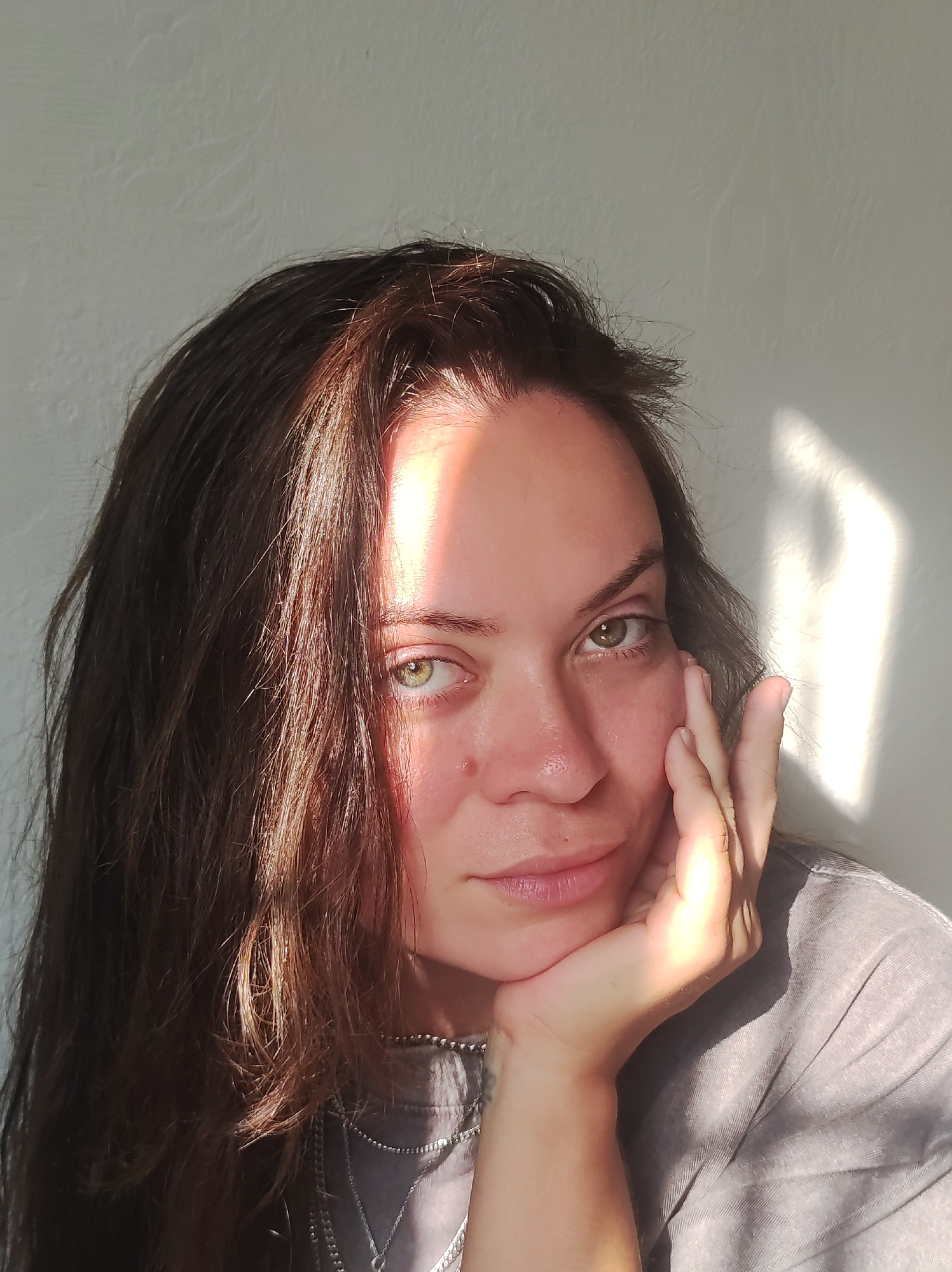 A woman is pictured in a close-up, head-and-shoulders shot against a textured, light-colored wall. The setting sun casts a strong beam of light across her face and hair, creating a dramatic contrast. Her right hand is resting on her chin, with her fingers splayed against her cheek. She has long, dark brown hair that falls around her shoulders and face. Her eyes are a striking green. She is wearing a grey t-shirt with a silver necklace. The image does not contain enough information to determine its location in Chaikovsky, Russia.