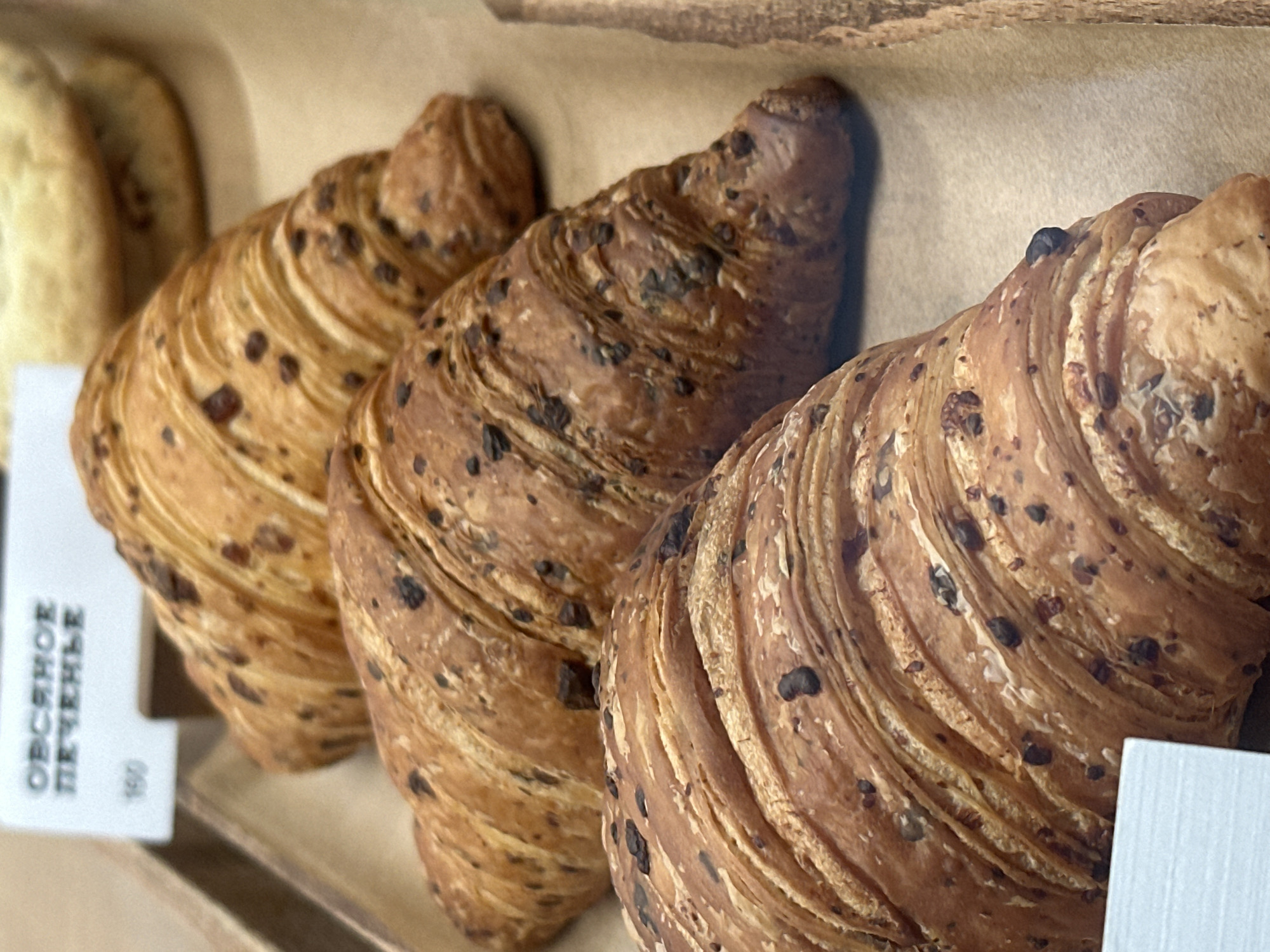 The image provides a close-up view of three golden-brown croissants, likely chocolate croissants, arranged diagonally on a display surface. The pastries exhibit a flaky, layered texture, characteristic of puff pastry, and are visibly studded with numerous dark chocolate pieces or chips. The croissant on the far right is in sharp focus, highlighting its intricate details, while the middle one is also well-defined. The leftmost croissant is slightly softer in focus but still clearly identifiable. Behind the leftmost croissant, a blurred, lighter-colored baked item, possibly a cookie or another pastry, is partially visible.
The baked goods are resting on a light brown surface, suggesting they are presented in a bakery, cafe, or store display case. In the bottom-left corner, a white label is partially visible. It features Cyrillic text that reads "ОВСЯНОЕ ПЕЧЕНЬЕ" (OVSYANOE PECHENYE), which translates to "Oatmeal Cookie." Below this text, the number "100" is also visible, likely indicating a price or weight. The presence of Cyrillic text confirms the location context of Kazan, Russia. The scene depicts an enticing display of fresh pastries ready for purchase. No people are visible, and the time of day or specific event cannot be determined from the image.