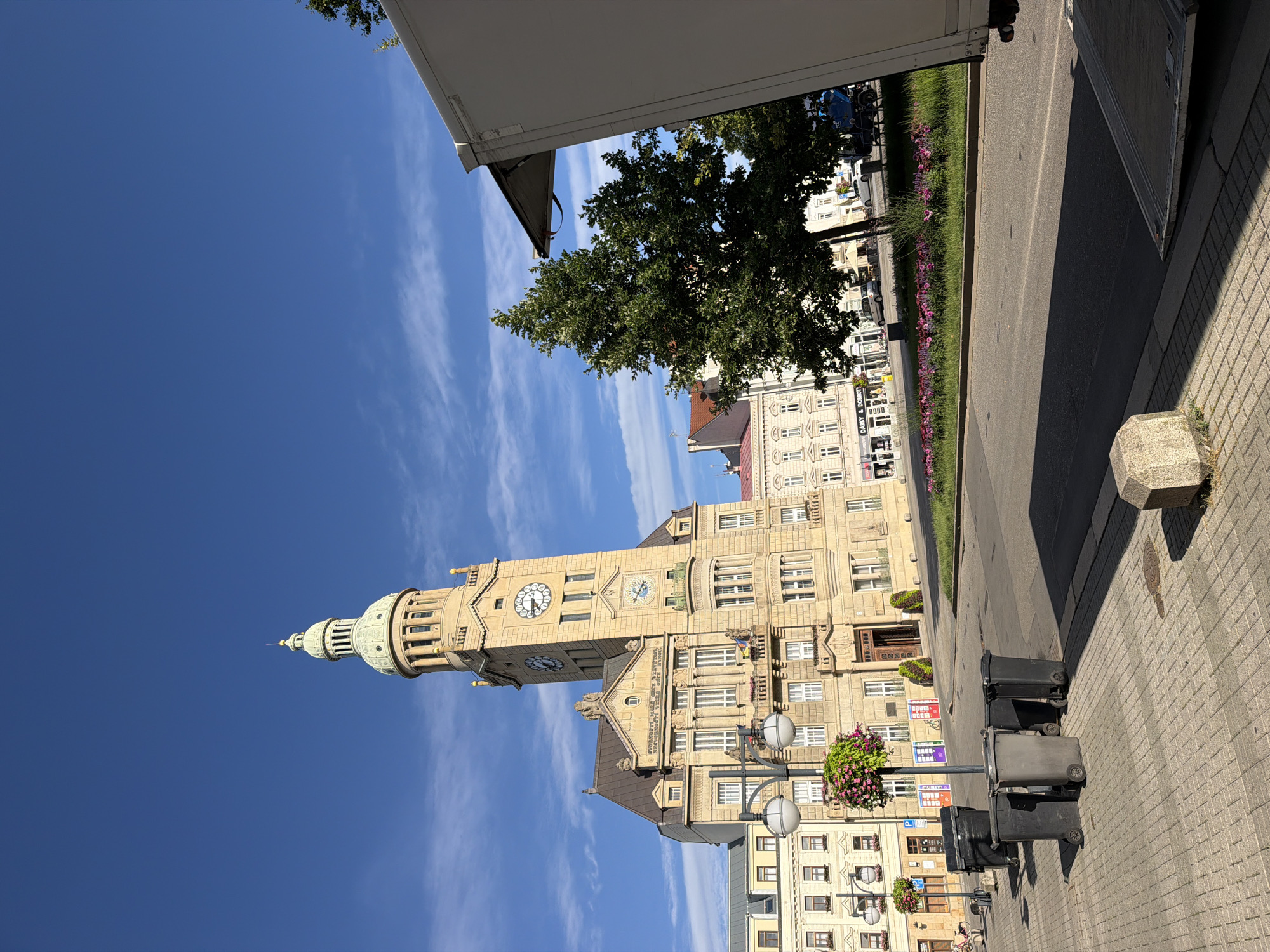 This is a photograph of a city square in Prostějov, Czechia, on a clear day. A tall, ornate building with a clock tower is the central architectural feature, rendered in a light-colored stone. The tower has a green, domed cupola. In the foreground, there is a paved sidewalk with several dark-colored trash bins. A street runs alongside the sidewalk, with a green median strip planted with flowers. A large, white trailer or truck is parked on the right side of the frame, partially obscuring a tree. The sky is a vibrant blue with scattered white clouds. There are no people visible in the immediate foreground, though some activity can be inferred on the street and in the background buildings, which appear to be shops.