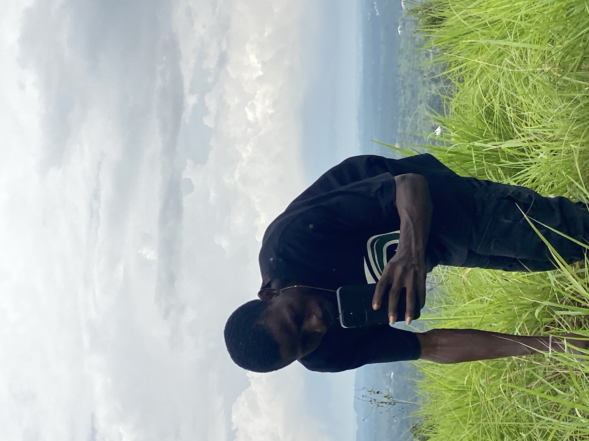 A young Black man is positioned in the foreground, standing in tall green grass on a hillside. He is wearing a black t-shirt and black pants, and is looking down at a black smartphone he is holding in his right hand. His left arm is bent, and his hand is resting on the grass. In the background, there is a lush, green landscape with rolling hills and a hazy sky filled with clouds. The setting appears to be outdoors in a rural or semi-rural area. It is daytime. The specific location is on a hillside in Nguzu Edda, Nigeria.