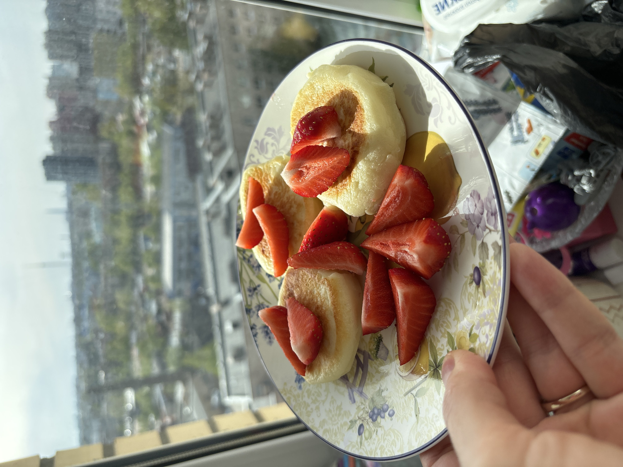 A plate of what appears to be Syrniki (a type of Russian fried cheese pancake) garnished with sliced strawberries is being held near a window. The plate is decorated with a floral pattern. A hand with a ring on the finger is holding the plate. Through the window, a blurred view of city buildings and a street scene is visible.