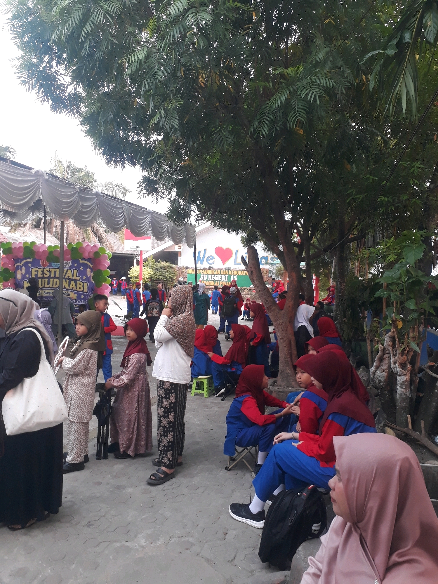 In Pangkalpinang, Indonesia, an outdoor event appears to be underway. A banner with "FESTIVAL MAULID NABI" is visible, along with balloons. Many children, some in uniform blue and red attire, are gathered. Adults are also present, some wearing hijabs. The setting seems to be a schoolyard or public space, with tents and trees providing shade. Some individuals are seated on the ground or on small chairs, while others are standing and observing. The ground is paved with interlocking concrete blocks. The overall atmosphere suggests a community gathering or celebration.