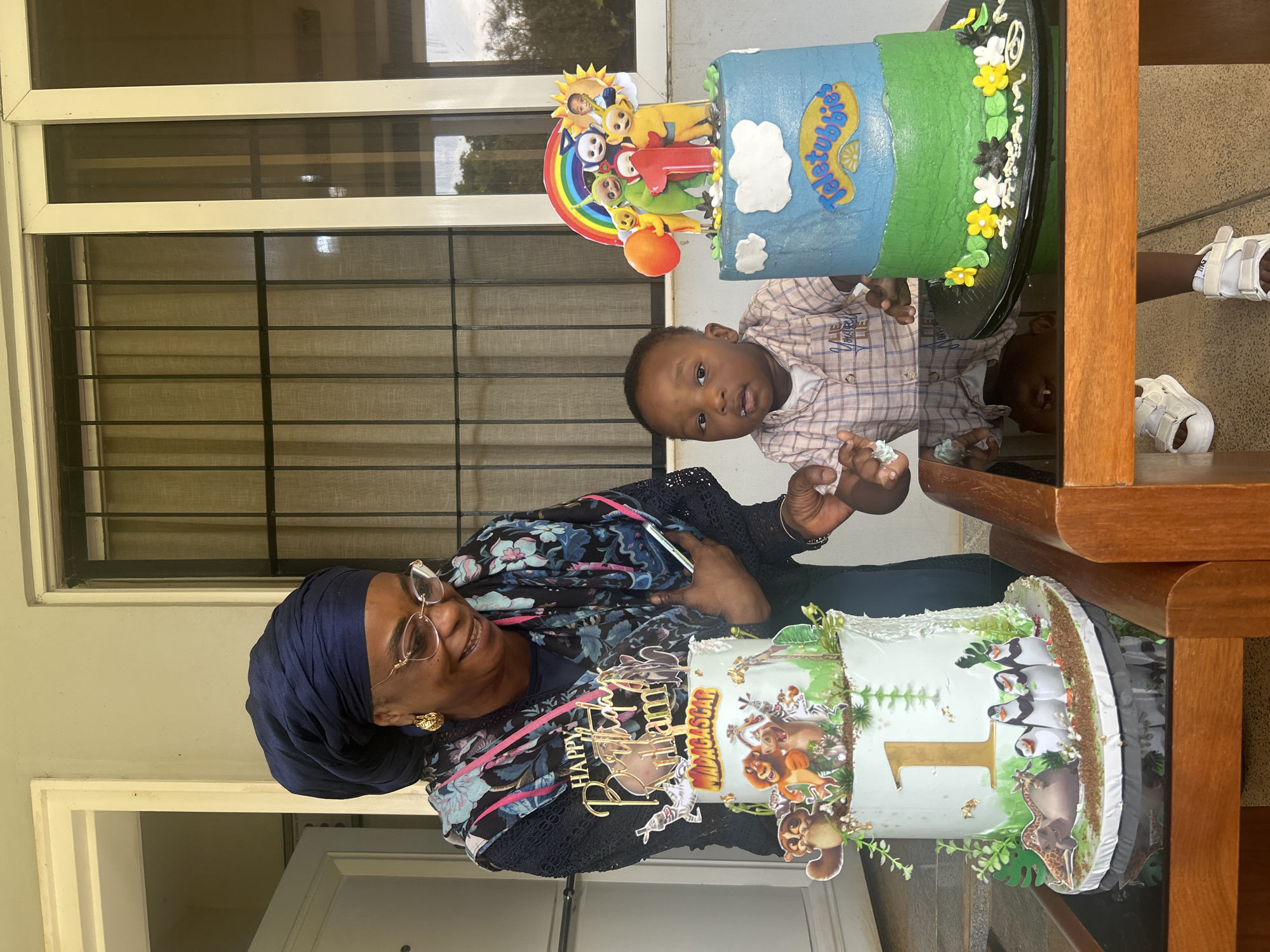 An elderly Black woman wearing a dark blue headscarf and glasses, with a colorful patterned scarf draped over her shoulders, smiles and looks down at a young Black child in a light brown checkered shirt. The woman is standing behind two birthday cakes. The cake on the left is a two-tiered cake decorated with characters from Madagascar, a golden number "1", and penguins. The cake on the right is a two-tiered cake with a Teletubbies theme, featuring characters from the show, a rainbow, and a sun. The child is seated in front of the Teletubbies cake, looking towards the camera. The scene appears to be outdoors in front of a building with large windows and a doorway. The setting is likely a residential area in Dutsen Kato, Nigeria, given the context provided.