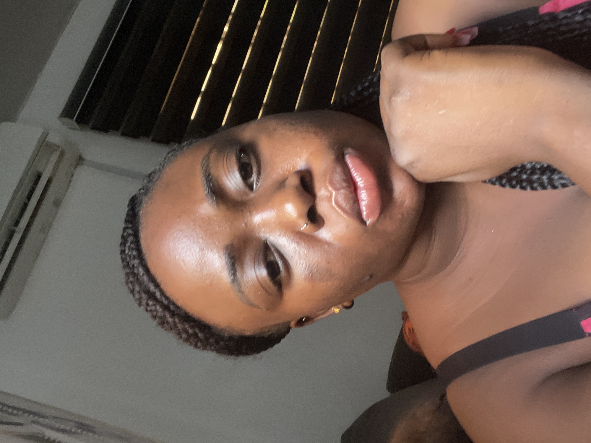 A close-up, selfie-style photo shows a person with dark skin and braided hair. The person's right cheek rests on their right hand. A nose ring is visible on their left nostril. They are wearing a black top with pink accents. In the background, out-of-focus, are horizontal blinds and a white, air-conditioning unit. Given the prompt's request for a location, it's in the city of Abuja, Nigeria, but no specifics can be deduced.