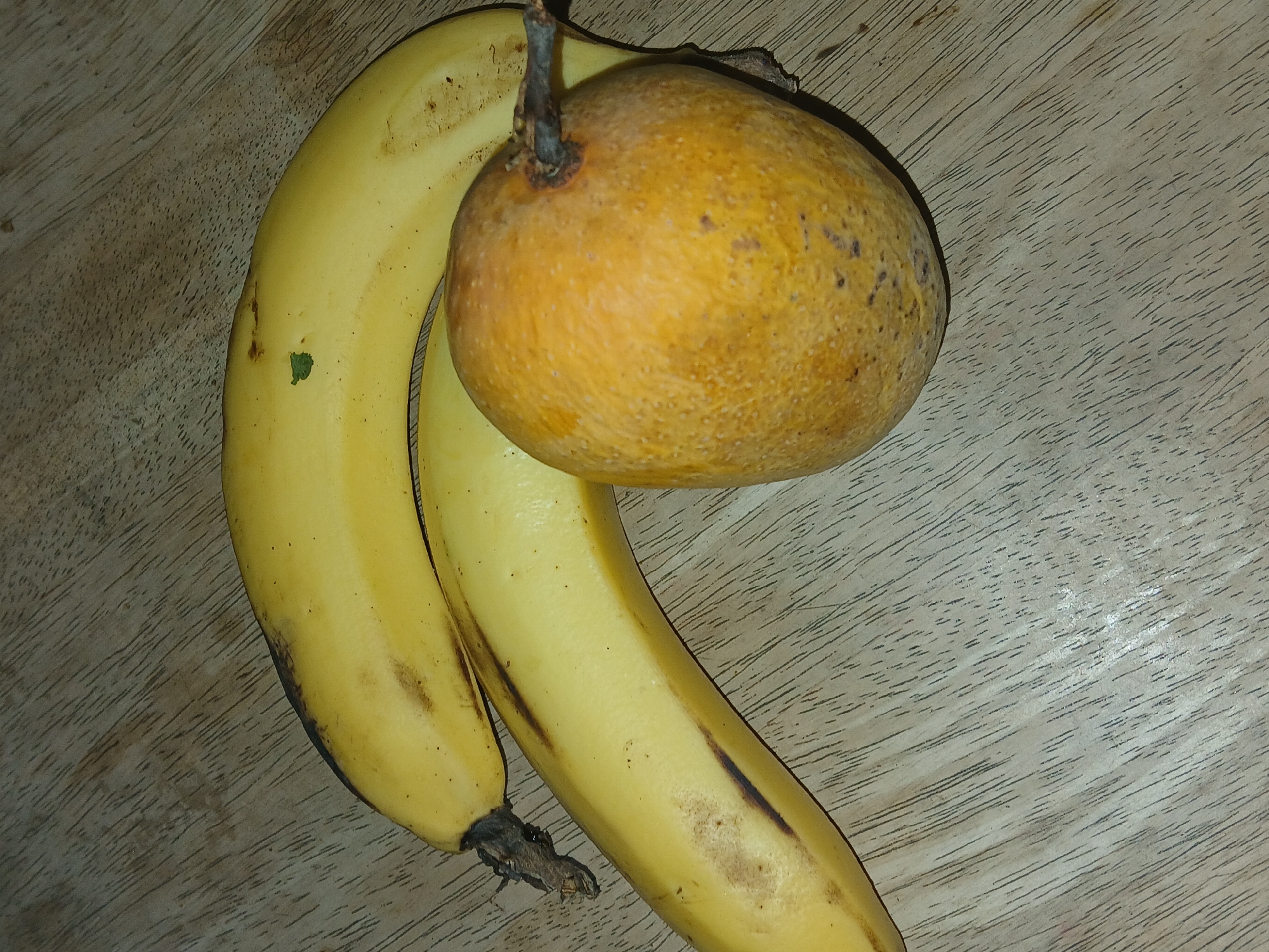 On a light brown, wood-grain surface lie two yellow bananas and a round, orange fruit. The bananas are curved and touching each other, with slight brown spots on their peels. The orange fruit, which has a stem, rests partially on the bananas. The surface appears to be a table or countertop. The location in Bengaluru, India is unknown.