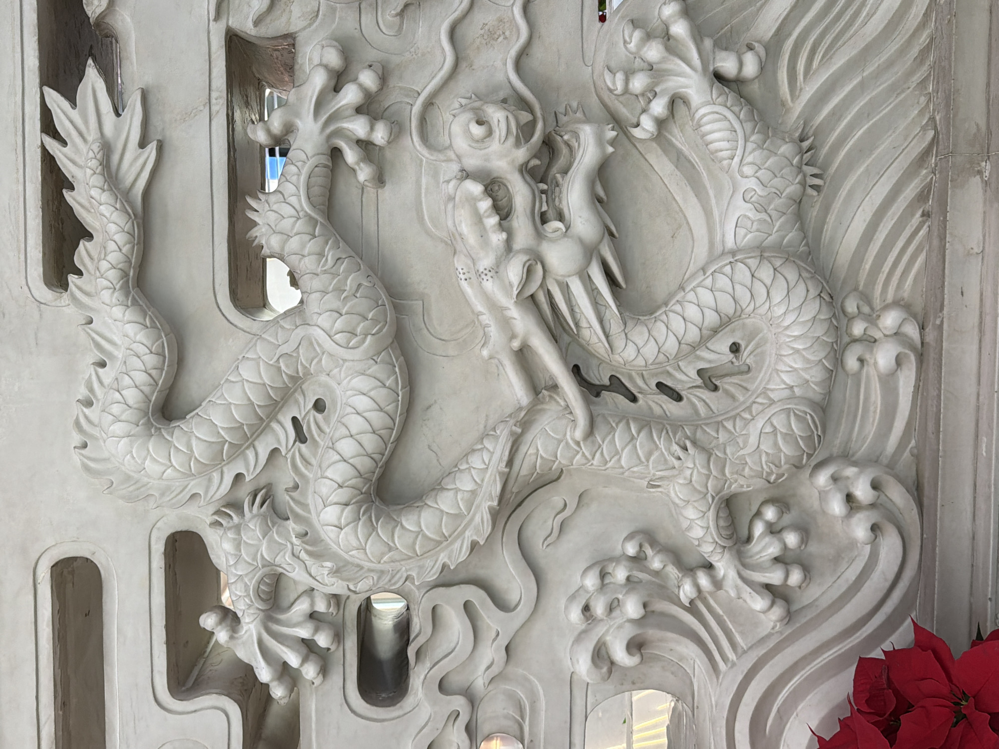 The image shows a white, bas-relief carving of two intertwined dragons. The carving features intricate detailing of the dragons' scales, claws, and faces. The background of the carving includes stylized clouds and waves. The carving appears to be part of a wall or panel, featuring rectangular openings or slots. Red poinsettias are visible in the lower right. The dragons' image appears to be on the location in the city of Shunyi District, China.